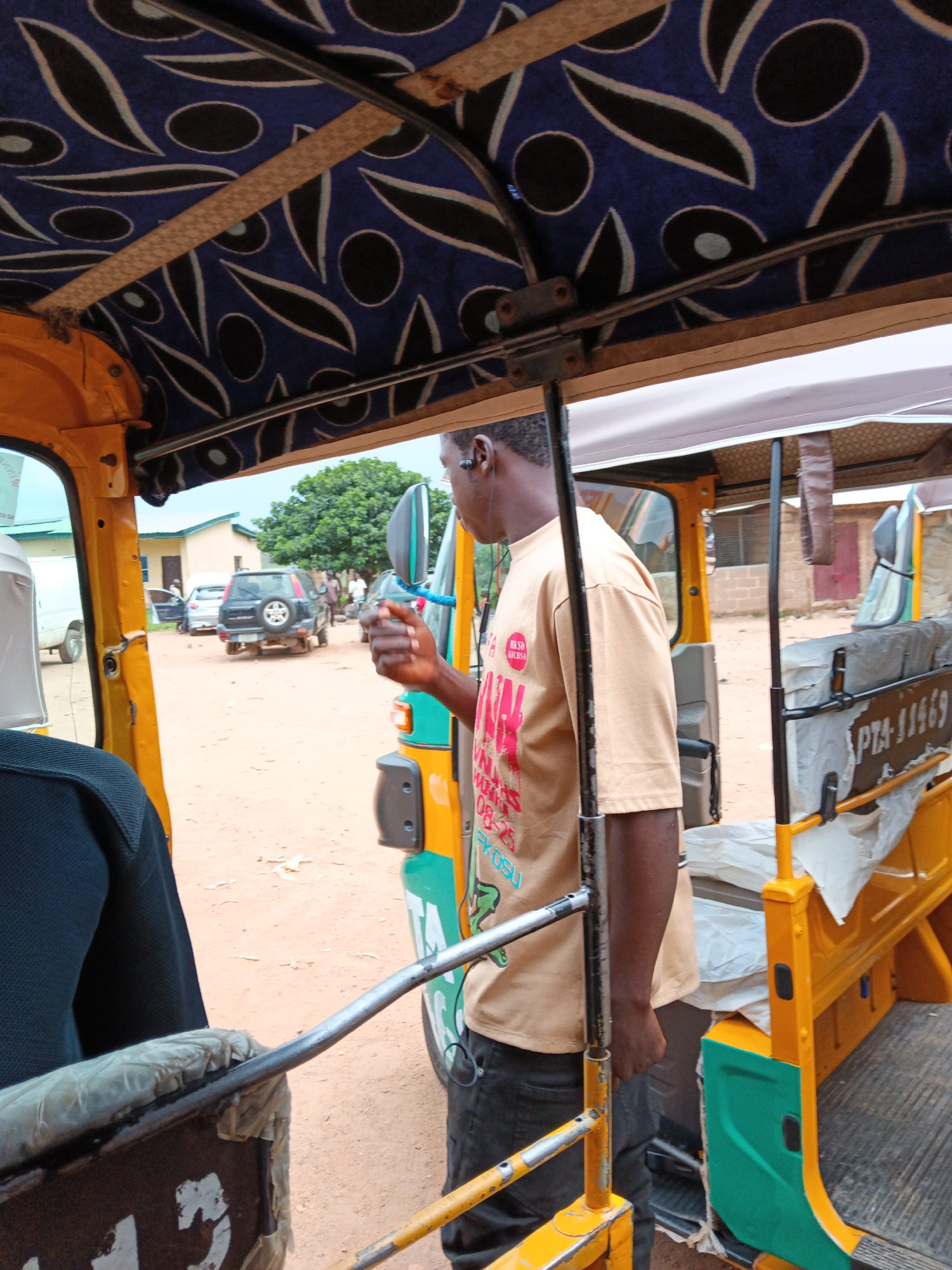 This media file shows the interior of a yellow auto-rickshaw, also known as a tuk-tuk. The roof lining of the auto-rickshaw is a dark blue fabric with a pattern of white and black oval shapes. A person, a young Black male, is visible in the foreground, wearing a light brown t-shirt with red text and graphics. He appears to be wearing headphones.
The scene outside the auto-rickshaw is a dusty, outdoor setting. In the background, there are buildings, some made of brick, and a large green tree. Several vehicles are parked in the area, including a dark SUV, a white van, and what appears to be another auto-rickshaw. The ground is a sandy, reddish-brown color, indicative of a dry, possibly arid environment. The lighting suggests it is daytime, with diffused light, implying it might be overcast or a cloudy day.
The person in the foreground is looking to the side, and his hand is raised slightly, holding what might be a phone or another small object. There is some visible text on the person's t-shirt, though it is partially obscured and difficult to read in full. The text appears to include "KOSU" and a date or series of numbers like "08-25". There is also text on the side of the auto-rickshaw, reading "PTA-11469". The overall impression is a snapshot of daily life in a location where auto-rickshaws are a common mode of transport.