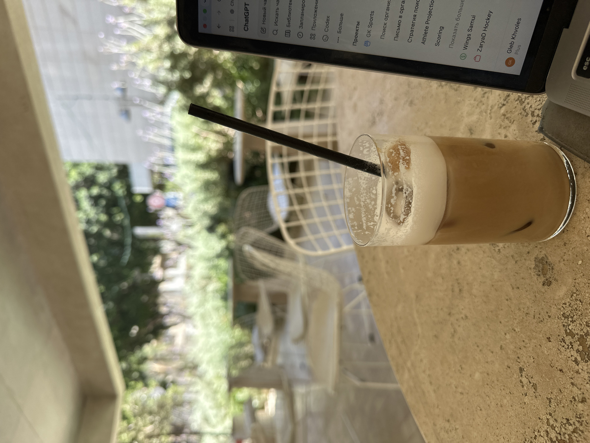 The image shows a close-up of a glass of iced coffee with a dark straw on a textured table. To the right, part of a laptop screen is visible, displaying text in Russian, including "ChatGPT", "Новый час" (New hour), and "Искать чат" (Search chat). The background is blurred, showing outdoor seating with chairs and tables, greenery, and what appears to be a building facade. The presence of outdoor seating, greenery, and the architectural style suggests a cafe or restaurant setting. Without specific landmarks or clear identifying signs, pinpointing the exact location within Tel Aviv is not possible from the image alone. The scene depicts a person working or relaxing at a cafe with their iced coffee and laptop.