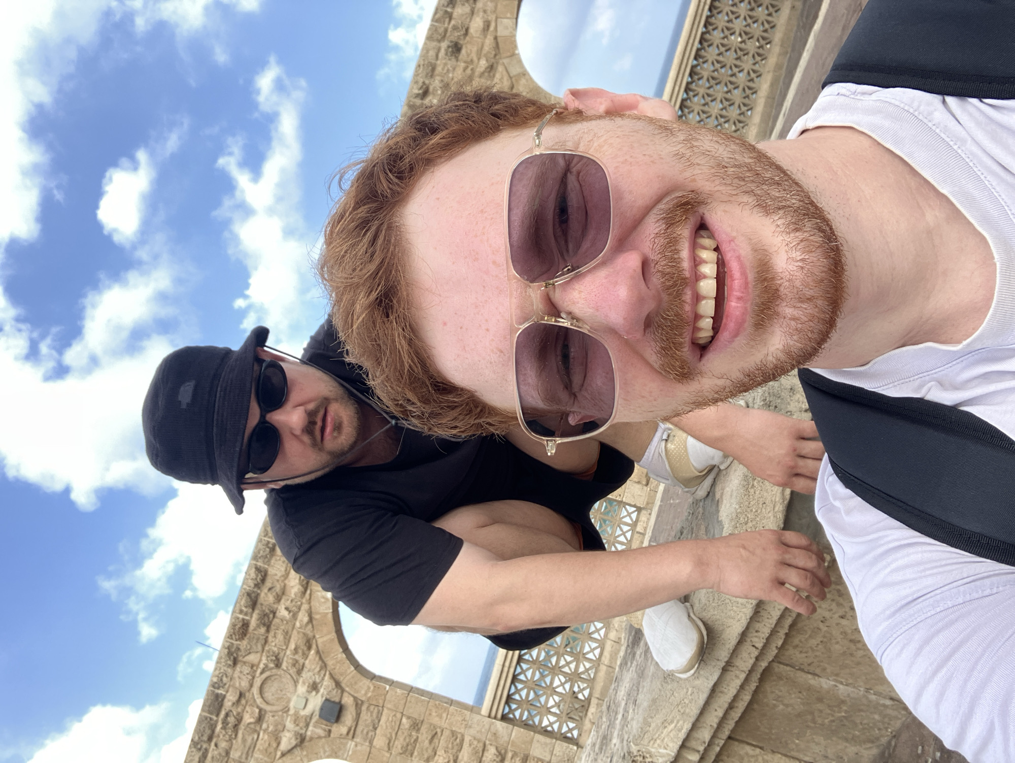 Two individuals are captured in a selfie outdoors, with a stone archway in the background and a cloudy blue sky above. The foreground is dominated by the face of a man with red hair and a beard, wearing gold-rimmed sunglasses and a white t-shirt, with a backpack strap visible. Behind him, another man wearing a black bucket hat and sunglasses squats down, dressed in a black t-shirt and shorts. The man in the foreground appears to be in the city of Tel Aviv, Israel, judging by the architectural style of the stone structures and the clear sky, consistent with a Mediterranean climate. The overall scene suggests a casual outing, possibly at a historical or scenic site within the city.