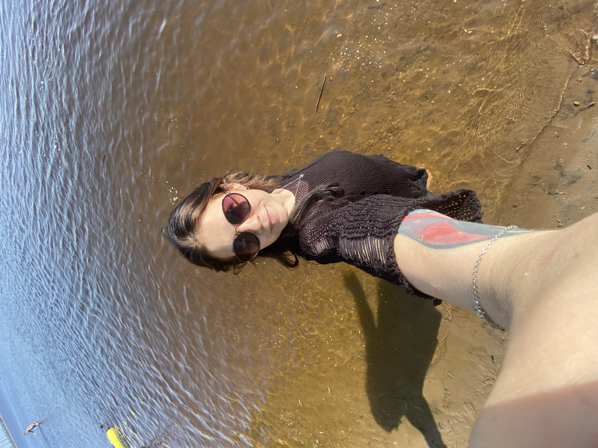 A selfie taken near the edge of a body of water, possibly the Kama River in Chaikovsky, Russia, judging by the color and appearance of the water. A woman wearing sunglasses and a dark, somewhat sheer, crocheted top is positioned in the foreground. She is partially in the water, which appears shallow and allows the sandy bottom to be visible. Her arm, adorned with a tattoo and a silver anklet, is extended toward the camera. The shoreline is visible on the right side, consisting of sand and small debris. In the background, the water extends towards the left, with the opposite bank faintly visible in the distance. There is a person swimming in the distance. The image is brightly lit, suggesting a sunny day.