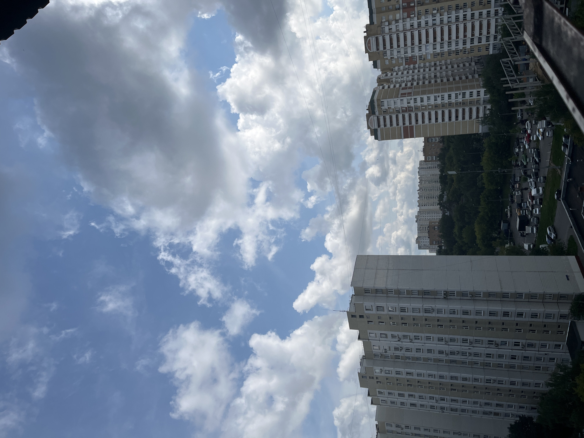 This media file depicts a high-angle view of a residential area in Moscow, Russia, on a cloudy day. The primary subjects are multiple apartment buildings of varying heights and architectural styles, as well as a street lined with parked cars. The buildings are predominantly light-colored with numerous windows and visible air conditioning units. The sky is filled with a mixture of dark, heavy clouds and patches of bright blue, suggesting a dynamic weather system. Thin power lines are visible crisscrossing the sky between the buildings.
The scene suggests a typical urban environment, likely during the daytime given the visibility and lighting. The presence of many parked cars indicates a residential neighborhood where people live. There is no discernible activity of people in the frame; the focus is on the static elements of the cityscape and the sky. The weather appears to be partly cloudy with a potential for rain or a recent shower, judging by the darker cloud formations. The location cues, such as the style of the apartment buildings and the mention of Moscow, Russia, help to contextualize the image. No visible text is present in the media file. The overall mood evoked is one of urban life under a changing sky.