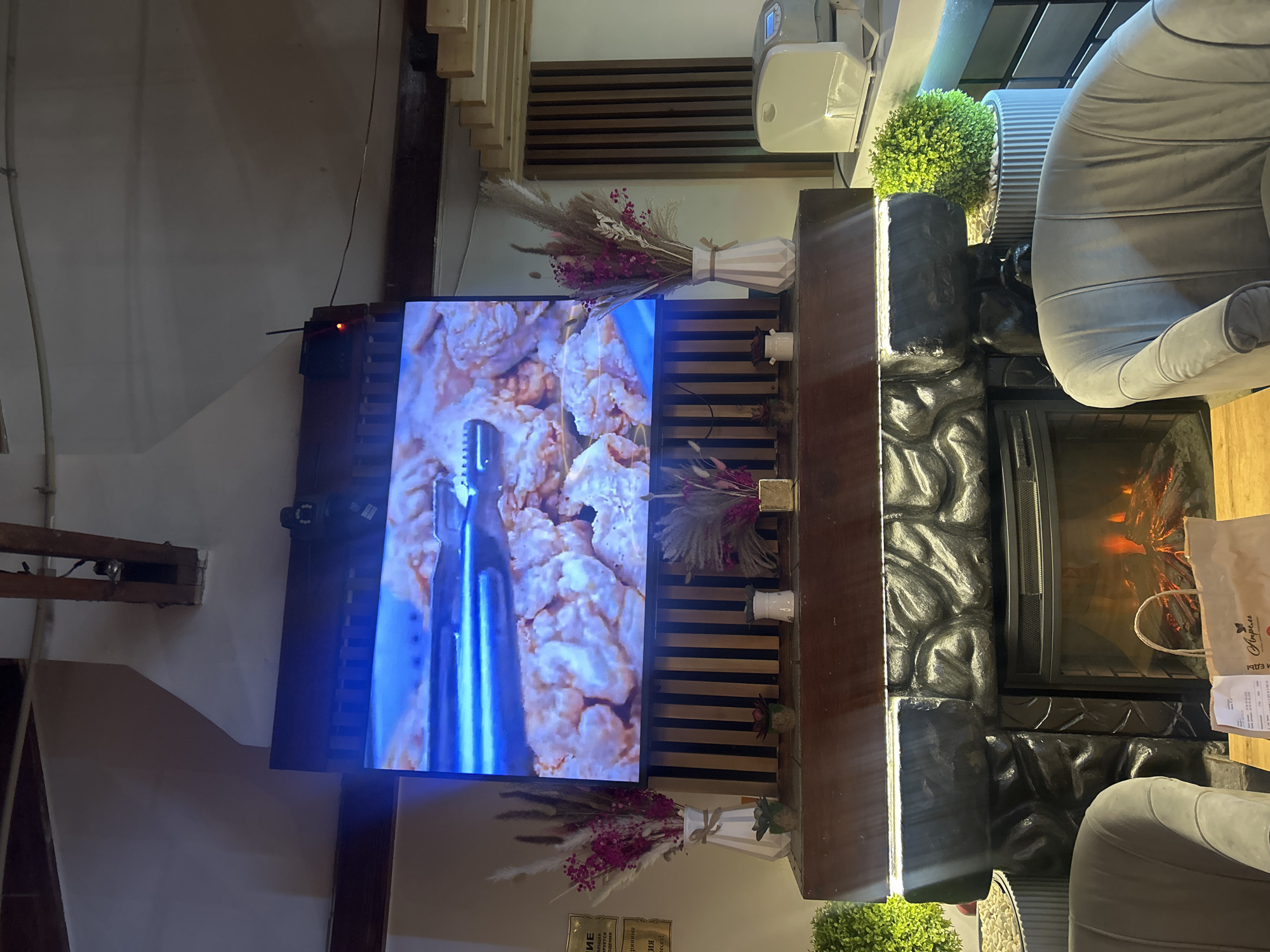 The image depicts a cozy cafe interior, possibly located in Almaty, Kazakhstan. A large flat-screen television is mounted above a decorative fireplace, displaying a close-up of fried chicken. The fireplace itself is made of dark, textured material and has an electric fire burning. On the mantelpiece, there are various decorative items, including vases with dried flowers and small potted plants. To the right of the fireplace, a white appliance, possibly a bread maker or a small oven, is visible. In the foreground, plush grey armchairs are arranged, suggesting a comfortable seating area. A paper shopping bag rests on a table in front of the armchairs. The overall ambiance suggests a relaxed dining or lounging space. The lighting is warm and inviting, with additional illumination coming from the television and accent lights under the mantelpiece. The walls are painted in a neutral tone, and the ceiling features exposed wooden beams.