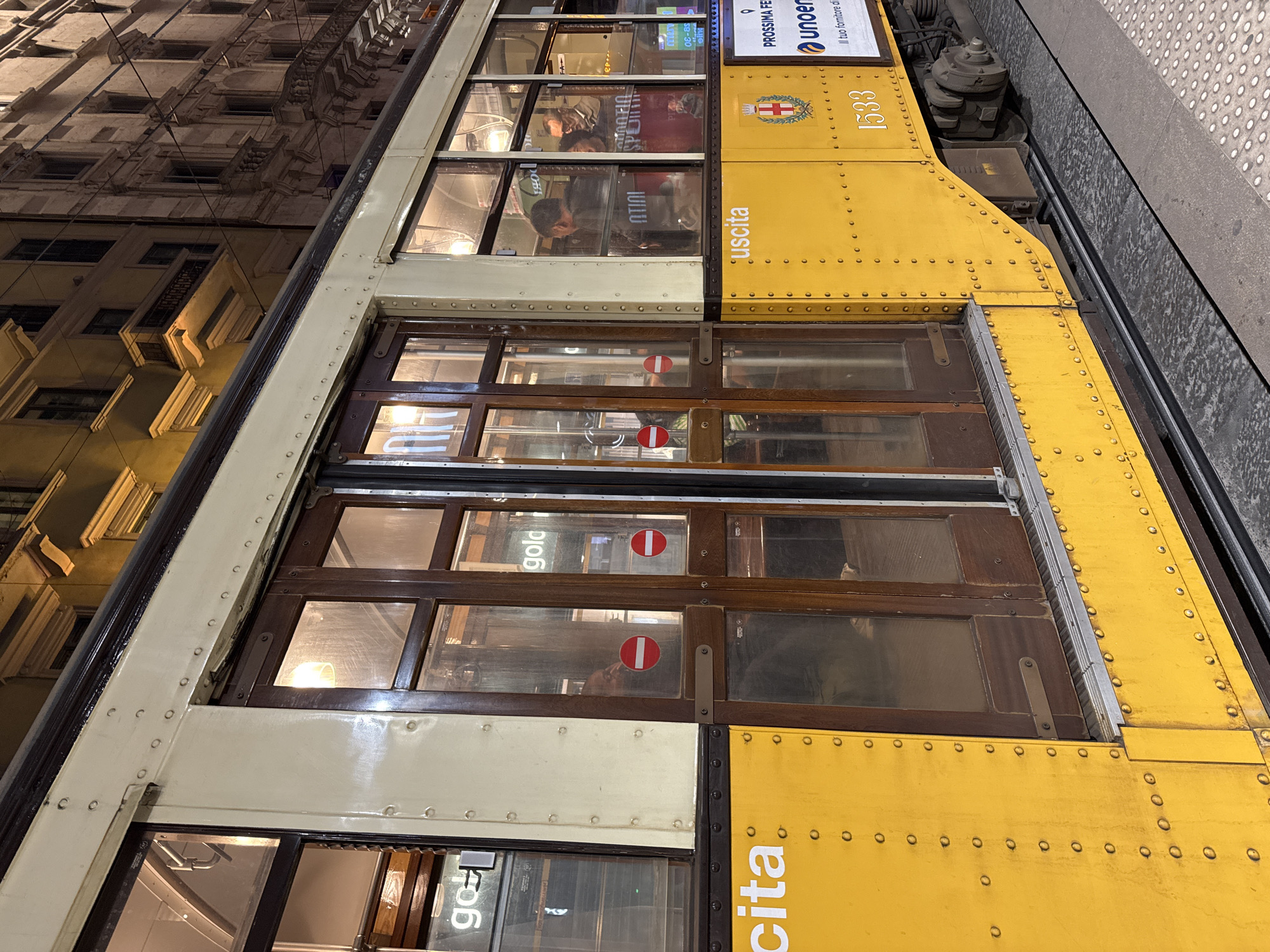 A vintage yellow and cream tram, identified by the number 1533 and a coat of arms, is captured at night in an urban setting, likely Milan, Italy. The tram features dark wooden window frames and doors, with numerous rivets visible on its metal body. Reflections of illuminated multi-story buildings with ornate architectural details and balconies are visible in the tram's windows and background.
Inside the tram, lights are on, and blurred reflections of several passengers can be seen. Two red circular "no entry" signs are present on the tram's glass doors. The word "uscita" (Italian for "exit") is prominently displayed in white text on the yellow side of the tram, indicating an exit point. Above the yellow section, a white sign partially reads "PROSSIMA FERMATA" (next stop), "unoer," and "Il tuo fornitore di." The tram appears to be either stopped at a platform or moving slowly, with a textured gray platform visible to its right. The night time setting is evident from the dark sky and the bright internal lighting of the tram.