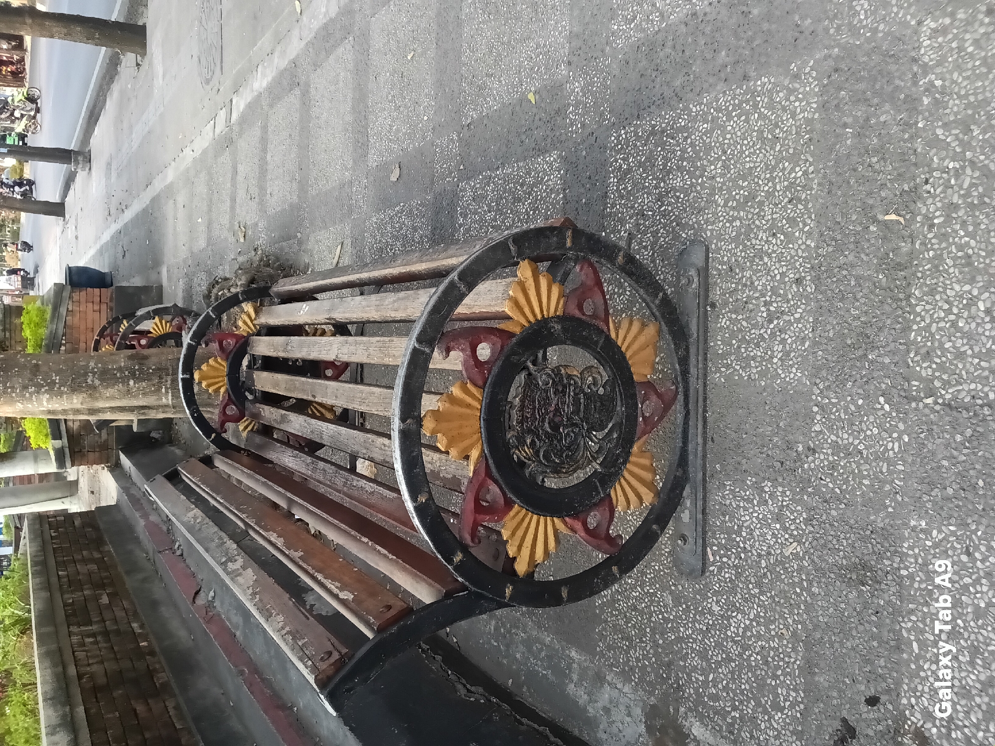 A weathered wooden bench with decorative black metal armrests featuring a sunburst design sits on a textured gray paved sidewalk. The bench is empty and unoccupied. The sidewalk is adjacent to a street where several motorcycles are visible in the background. A brick wall and some greenery are present to the left of the bench. The environment appears to be an outdoor urban setting. The timestamp "Galaxy Tab A9" is visible in the lower left corner of the image.
