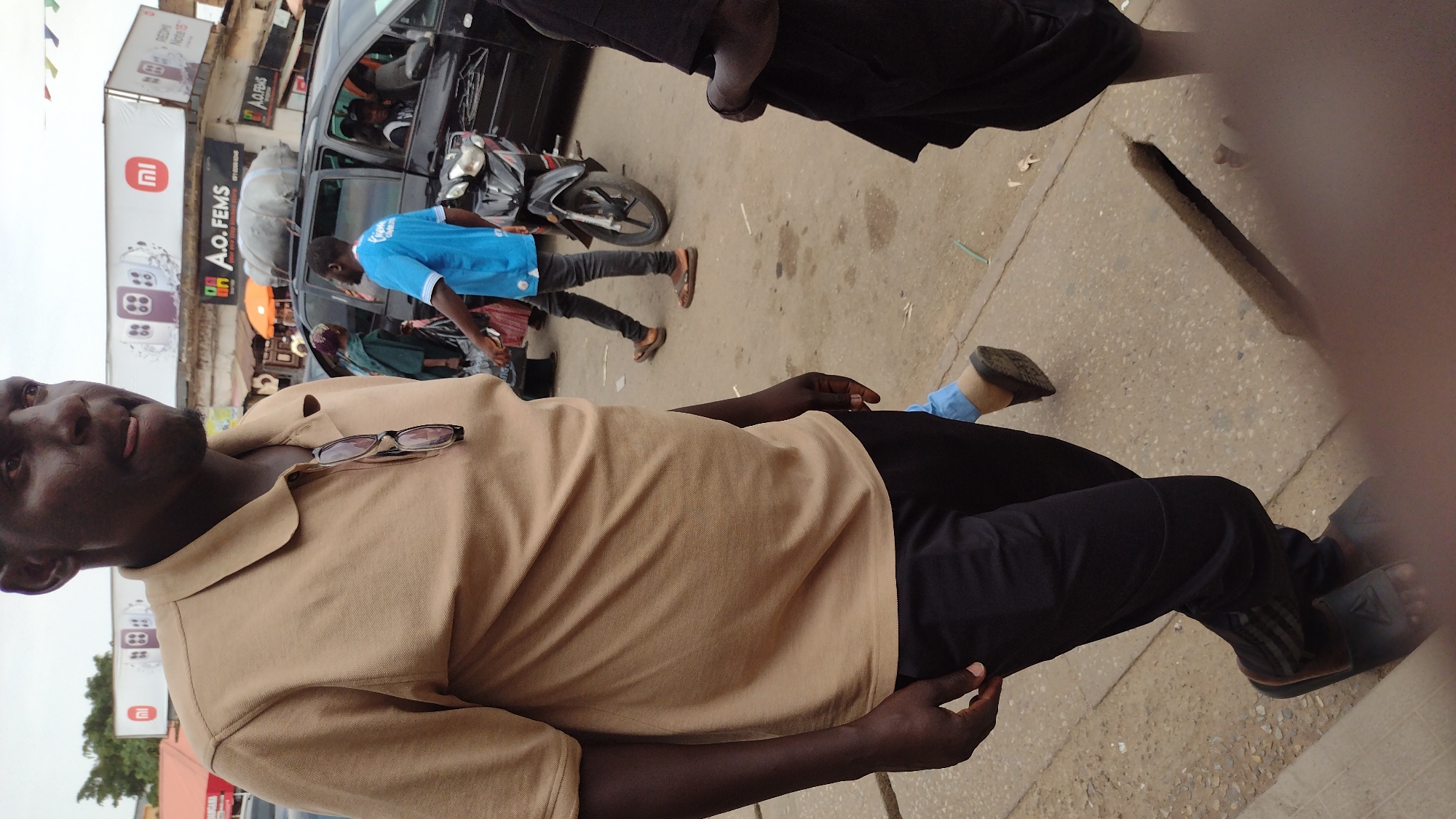 The image captures a street scene in Jos, Nigeria. A man in a tan shirt and black pants stands in the foreground, with glasses resting on his chest. Behind him, a man in a blue shirt walks past a black car, in front of an A.O. FEMS store with a Mi advertisement. A motorcycle is parked nearby. The background shows a busy street with other pedestrians and commercial signage.