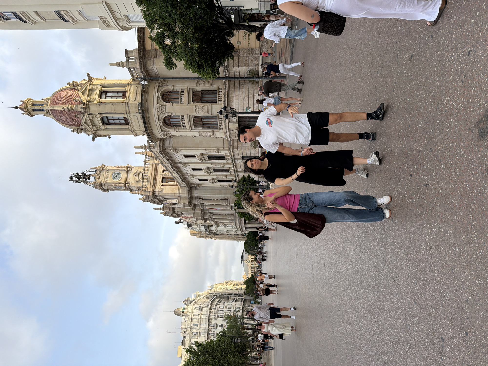 In the city of Valencia, Spain, on a wide, paved plaza, a group of people are walking under a cloudy sky. In the foreground, a young woman with blonde, curly hair, wearing a pink tank top and blue jeans, walks with her arm linked with a man. The man is wearing a white t-shirt with a graphic design, black shorts, and black socks with sandals. To their left, a woman in a black dress walks, her right hand in the man's left. In the mid-ground, other pedestrians are seen walking in both directions. Behind the pedestrians, grand, ornate buildings with classical architectural features and domed roofs line the plaza. One building features a prominent clock tower. Trees are visible along the sides of the buildings.