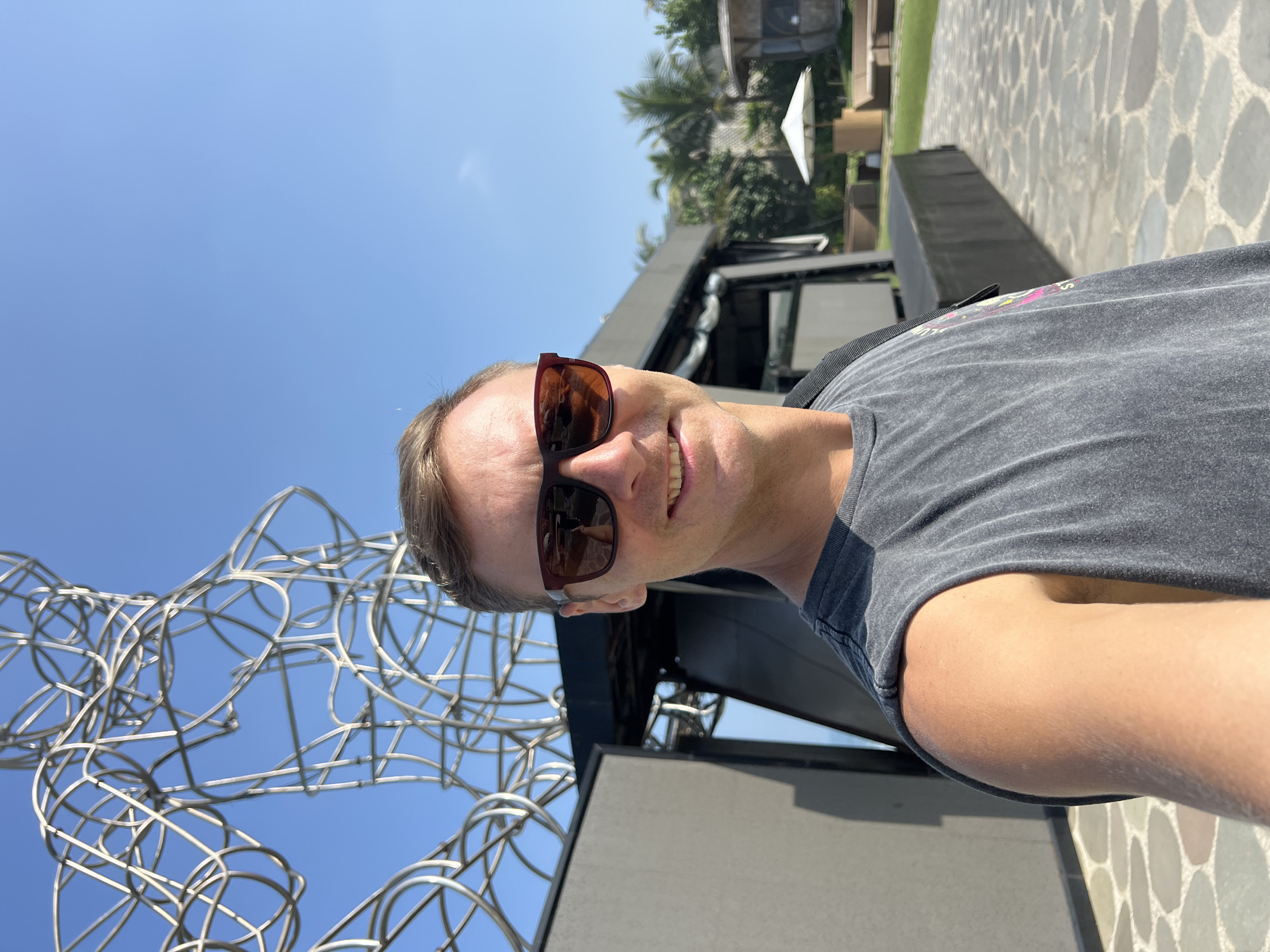 The image is a selfie taken outdoors on a sunny day with a clear blue sky. A man wearing sunglasses and a dark grey sleeveless t-shirt is in the foreground, smiling at the camera. Behind him, a large, abstract metal sculpture is visible against the sky. To the right of the sculpture, there is a modern building structure with dark walls and a black roof. Further in the background, there is a lawn area with outdoor furniture, including lounge chairs and a white umbrella. Palm trees are also visible in the distance. The ground in front of the man is paved with irregularly shaped stones. The image is a candid shot, likely taken during a vacation or outing. The location is not specified in Kediri, Indonesia.