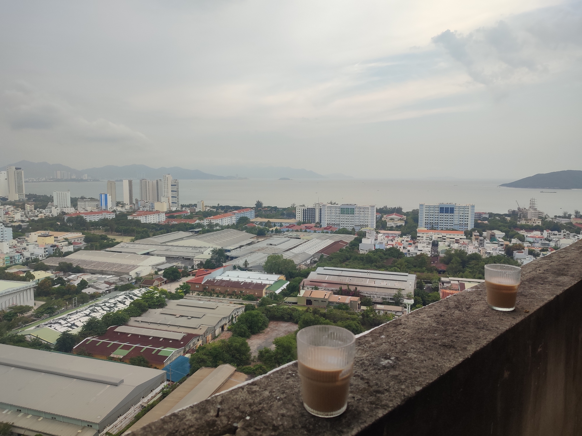 The image shows a cityscape with a cloudy sky and an ocean in the background. In the foreground, two glasses containing a brown liquid, possibly coffee or tea with milk, are placed on a concrete ledge. The city below features a mix of residential buildings, industrial structures with large roofs, and patches of greenery. In the distance, tall buildings are visible along the coastline, and mountains are discernible further out across the water. The image appears to have been taken in Nha Trang, Vietnam, given the recognizable cityscape and geographical features.