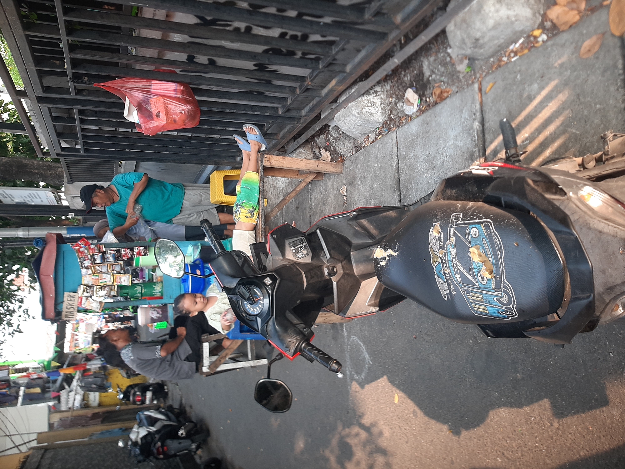 In the city of Central Jakarta, Indonesia, an outdoor scene unfolds with a focus on a street-level market or roadside vendor area. A dark-colored motorcycle is parked in the foreground, its seat bearing a sticker depicting a blue stylized vehicle, possibly a bus, with text above and below it. To the left of the motorcycle, a section of a sidewalk is visible, with shadows cast from overhead trees.
Behind the motorcycle, a young child with short, dark hair is seated and looking down, holding what appears to be a bottle. Next to the child, a man in a grey shirt and black pants sits on a stool. Further back, a small blue stall with a red roof serves as a vendor, displaying various packaged goods and a prominent sign reading "MASKER."
To the right of the stall, a man in a turquoise shirt and grey shorts stands and gestures with a bottle. He is positioned near a black metal fence. A young person, possibly a child, is lying on a wooden plank supported by wooden legs, with their legs extended towards the fence. They are wearing colorful shorts and blue flip-flops. A red plastic bag filled with items is hanging from the fence. Several scooters are parked in the background, and the overall setting suggests a busy, informal commercial area. The ground is paved, and there are scattered leaves and debris on the sidewalk.