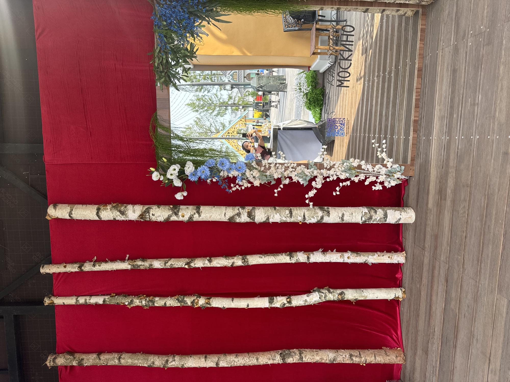 The image shows an outdoor setting in Moscow, Russia, with a red fabric backdrop and four vertical birch logs. In the foreground, a wooden deck leads towards an open doorway framed by greenery and white flowers. Through the doorway, a person is visible, holding a phone and appearing to take a selfie, with their reflection visible in the reflective surface of what appears to be a screen or glass. The background suggests a public space with buildings and trees. A QR code is visible on a white surface in the doorway, and the text "МОСКИТО" is printed on the floor in front of the doorway. The overall scene appears to be an outdoor event or installation.