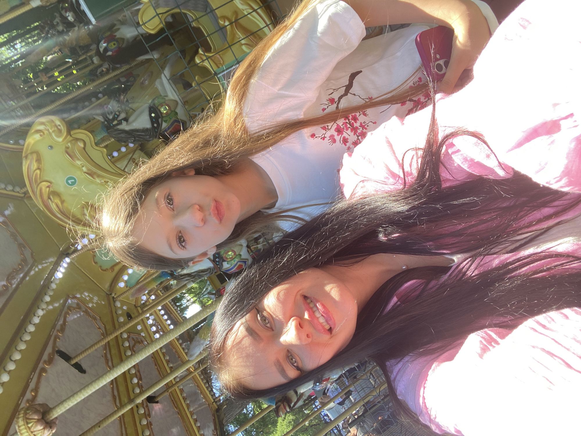 The image is a selfie taken by two females on a carousel. The person in the foreground has dark hair and is smiling. She is wearing a pink top. The person behind her has long, light brown hair and is puckering her lips. She is wearing a white t-shirt with a floral print. The carousel is decorated in gold and appears to be outdoors, with trees visible in the background. There is no discernible information in the image that would indicate the location is the city of Perm, Russia.