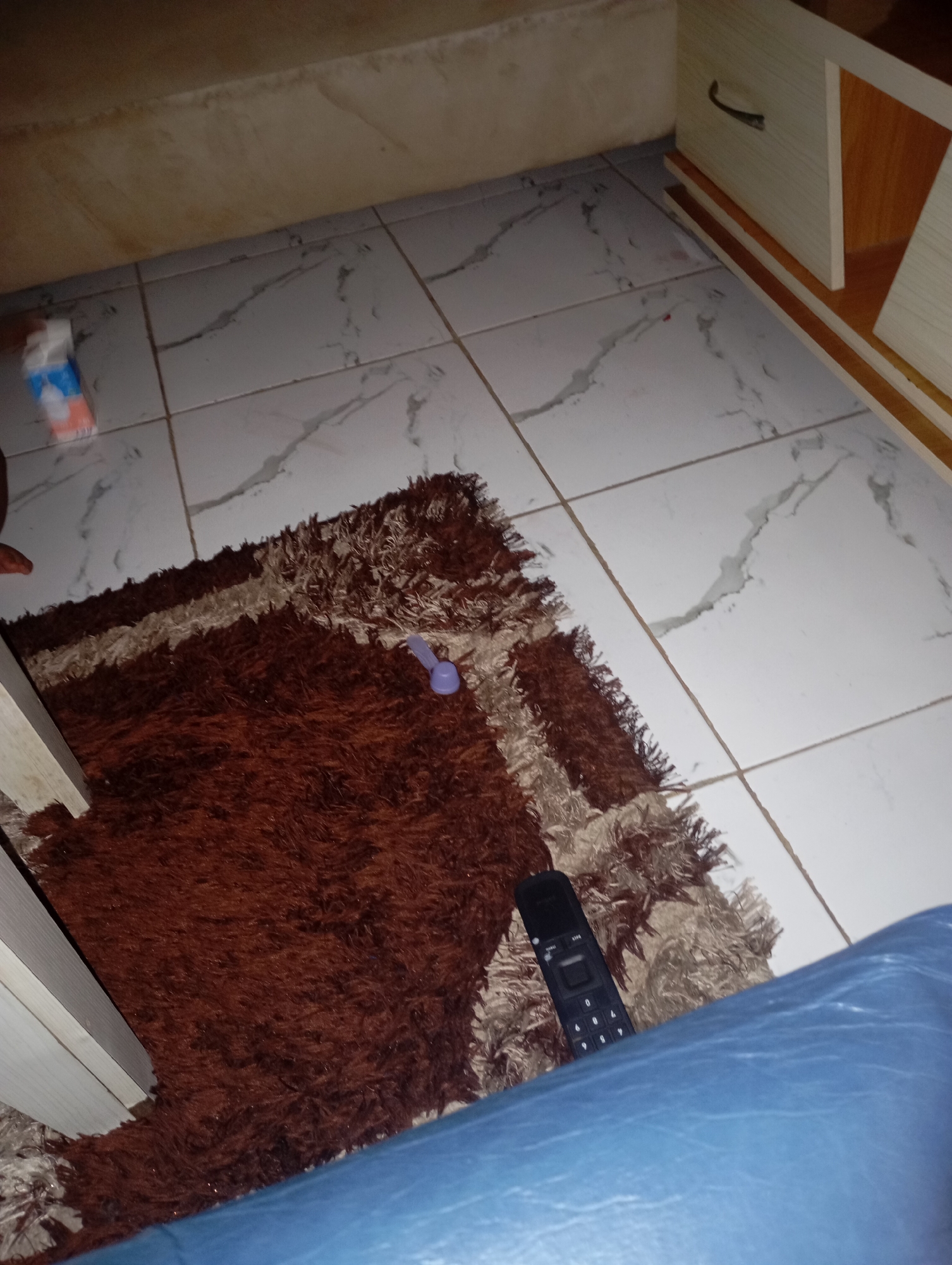 The image depicts an indoor scene, likely a room in a residential dwelling in New Stadium, Nigeria. The flooring appears to be tiled with a white and grey marbled pattern. A plush, shaggy rug with dark brown and lighter brown sections is prominently featured in the foreground, partially covering the tiled floor. To the left of the rug, a portion of a light-colored wooden piece of furniture is visible.
On the rug, a small, light purple object, possibly a toy or decorative item, is present. Further to the right on the rug, a black remote control with buttons is lying down. In the upper left corner of the image, a small, white object with blue and orange accents, resembling a small package or toy, is visible on the tiled floor. In the background, a wooden cabinet or drawer unit with a metallic handle is partially visible on the right side of the frame. The overall lighting suggests an indoor environment with some ambient light. No individuals are clearly visible in the frame, though a person's leg, clad in blue fabric, is in the bottom right corner, suggesting someone is present in the scene.