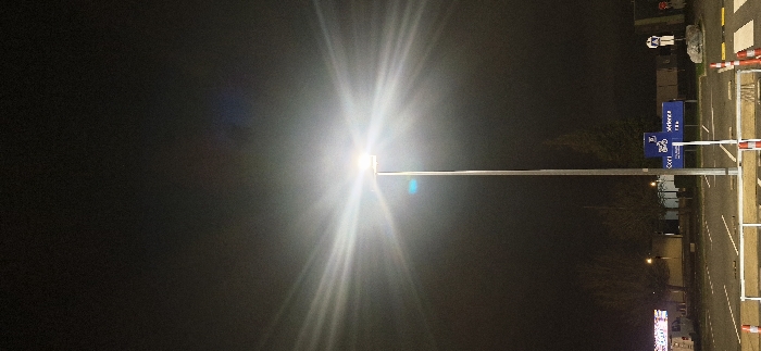 At night, a bright overhead light dominates the scene, its rays stretching across the dark background. To the right, a street with signage, barriers, and a sidewalk are visible. The sign displays information, including a bicycle icon. A crosswalk is marked with white lines. Trees and other structures are dimly illuminated along the street.