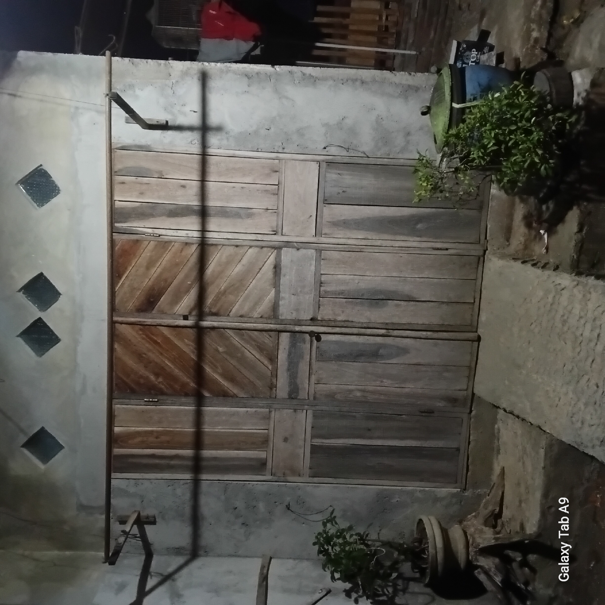 The image depicts an outdoor scene at night, likely in Trenggalek, Indonesia, showing a weathered wooden double door set into a concrete wall. Above the doors, diamond-shaped ventilation grates are visible in the wall. To the left of the doors, a small potted plant sits on a rustic base. To the right of the doors, another potted plant is situated next to a large blue container with a green lid, and a box labeled "TOP Coffee." Clothes, including red and black garments, hang on a line to the far right, behind a wooden fence. The ground is a rough concrete surface. The lighting suggests an artificial light source, casting shadows on the wall. The overall impression is a utilitarian exterior, possibly of a home or a small business. The text "Galaxy Tab A9" is visible in the bottom left corner, indicating the device used to capture the image.
