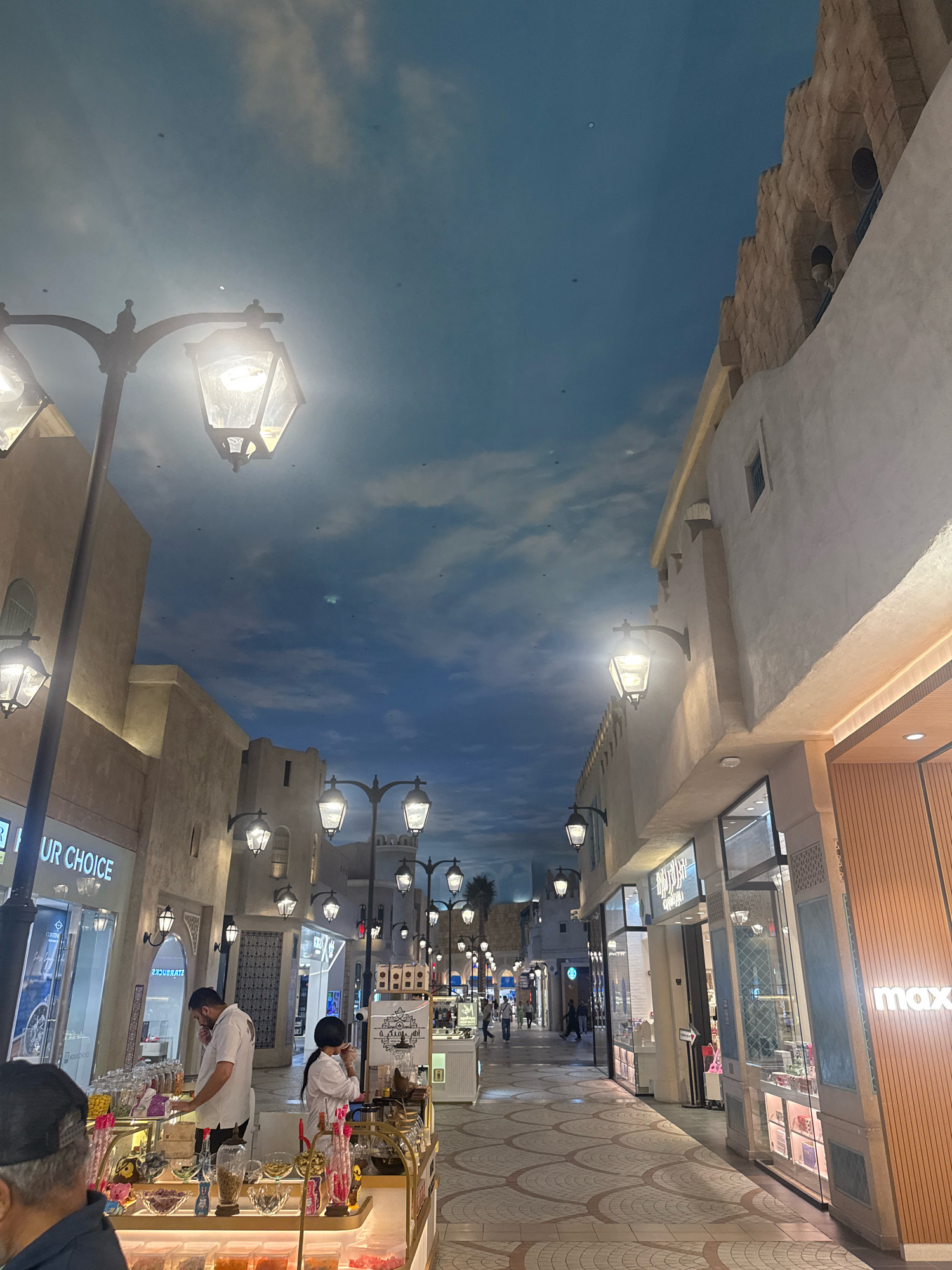 The image depicts an indoor marketplace in Dubai, United Arab Emirates, designed to resemble a Mediterranean or Middle Eastern streetscape with stucco-like buildings, arched windows, and decorative stonework. The ceiling is a large LED screen displaying a cloudy sky, creating an illusion of being outdoors. Traditional street lamps line the walkway, casting a warm glow.
In the foreground, a candy stall is visible with various colorful candies displayed in glass containers. Two individuals are interacting at the stall: a man in a white shirt appears to be assisting a woman in a white top who is holding a cup to her mouth. Another person, partially visible, wears a black baseball cap.
Further down the walkway, shops with glass storefronts are open. One shop displays the logo "max." Other signs in Arabic script are visible on the shop fronts. Several people are walking along the pathway, suggesting a public commercial space. The flooring is paved with a patterned tile design. The overall atmosphere is that of a well-lit, simulated outdoor shopping environment.