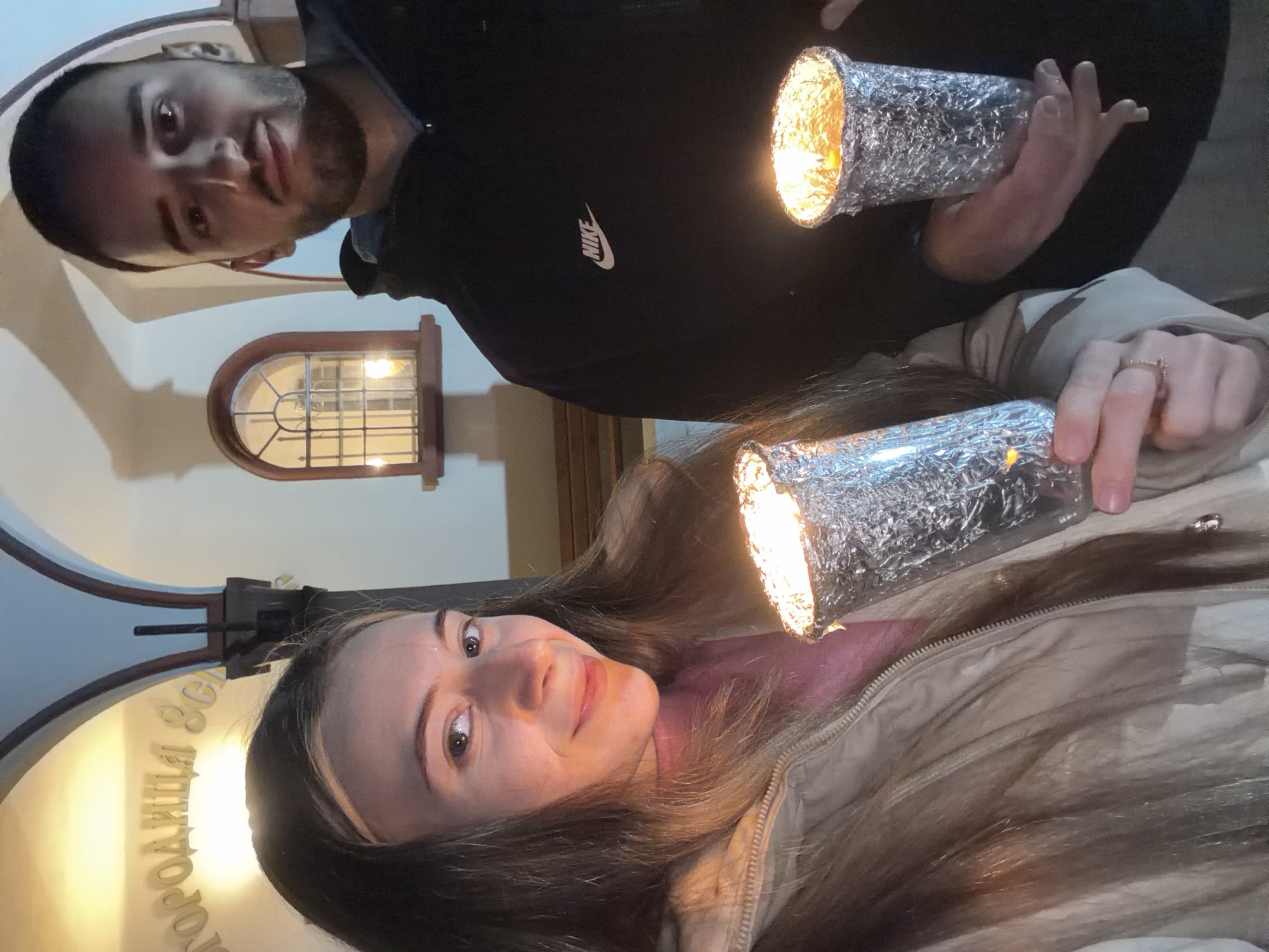 Two individuals are posing for a selfie inside a building in Milevo, Bulgaria. The woman has brown hair and is wearing a light brown jacket over a pink sweater. She is holding a foil-covered object that is emanating light. She is positioned in the foreground. The man is wearing a black Nike sweatshirt and is also holding a similarly wrapped, illuminated object. A small window is visible in the background. An archway with Bulgarian Cyrillic text above it suggests that the building is a business located within the city.