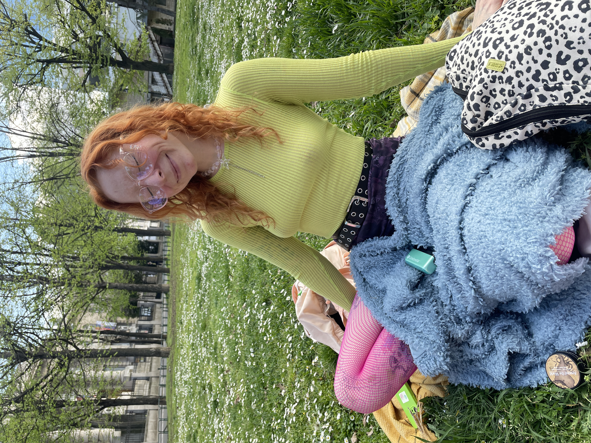 A young woman with reddish-orange hair and glasses sits on a grassy field covered with small white flowers. She wears a light green, long-sleeved shirt, a dark purple skirt with a black belt, and pink fishnet stockings. A blue furry blanket, a white and black leopard-print bag, and other assorted items surround her on what appears to be a blanket or picnic setup.
In the background, there are buildings and trees, indicating an urban setting. While it's difficult to pinpoint the exact location without more information, the scene suggests a park or green space within a city. Therefore it is difficult to specifically point out the location as the City of Palilula Urban Municipality, Serbia.