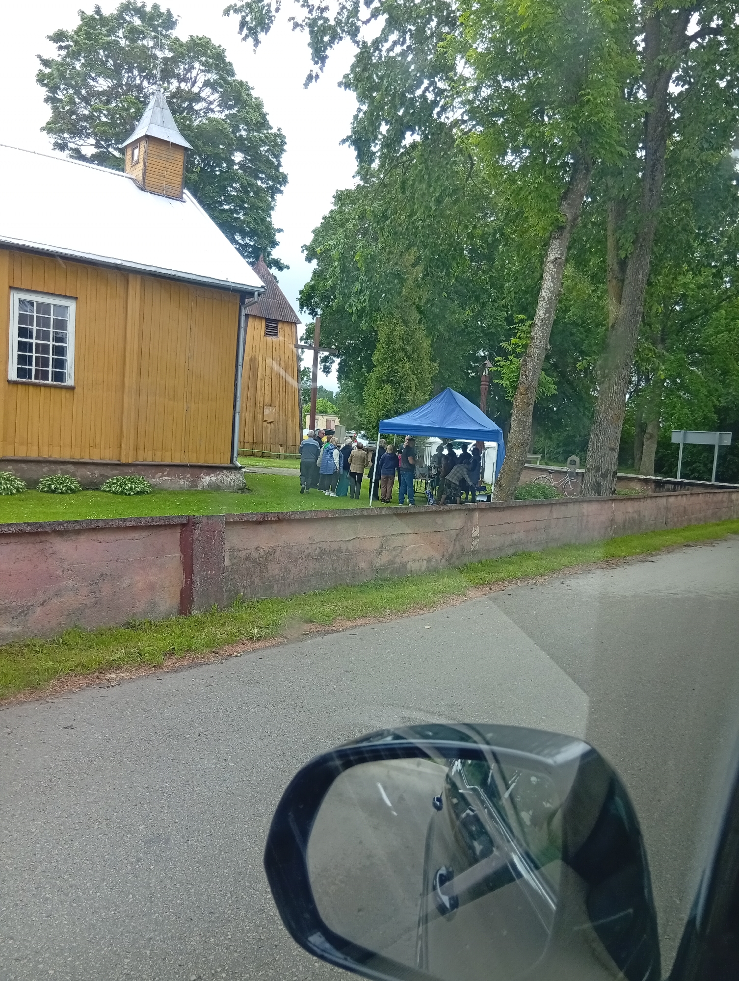 The image shows an outdoor scene in the village of Varputėnai, Lithuania. A yellow wooden building, possibly a church due to its architecture and steeple, is visible on the left. In front of the building, a group of people are gathered under a blue tent. Some individuals are standing and conversing, while others appear to be arranging items or engaged in activities beneath the tent. A low, reddish-brown wall runs along the perimeter of the property, separating it from a paved road in the foreground. Lush green trees and foliage surround the area, indicating a natural, possibly rural setting. A car's side mirror is prominently featured in the bottom right corner of the image, suggesting the photo was taken from inside a vehicle.