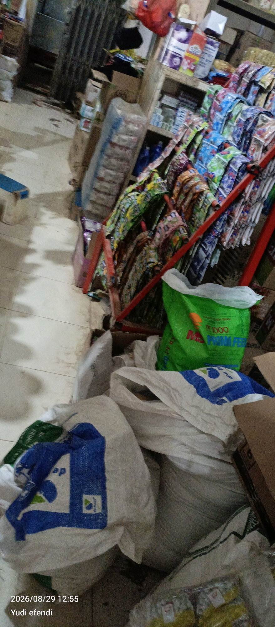 The image captures a cluttered interior scene, likely a small shop or storage area, located in Rawajitu Selatan, Indonesia. The foreground is dominated by piles of large, white sacks, some of which bear blue markings, suggesting they might contain goods. One sack in the lower left corner has a blue pattern and the text "ep" is partially visible. To the right of these sacks, a bright green sack with orange lettering is prominent. In the background, shelves are stocked with various packaged goods, many appearing to be small, colorful packets arranged in rows. A red metal rack is visible, supporting some of these shelves. The floor appears to be tiled. The overall impression is one of disarray and a busy commercial environment. The timestamp "2026/08/29 12:55" and the name "Yudi efendi" are superimposed on the lower left corner.