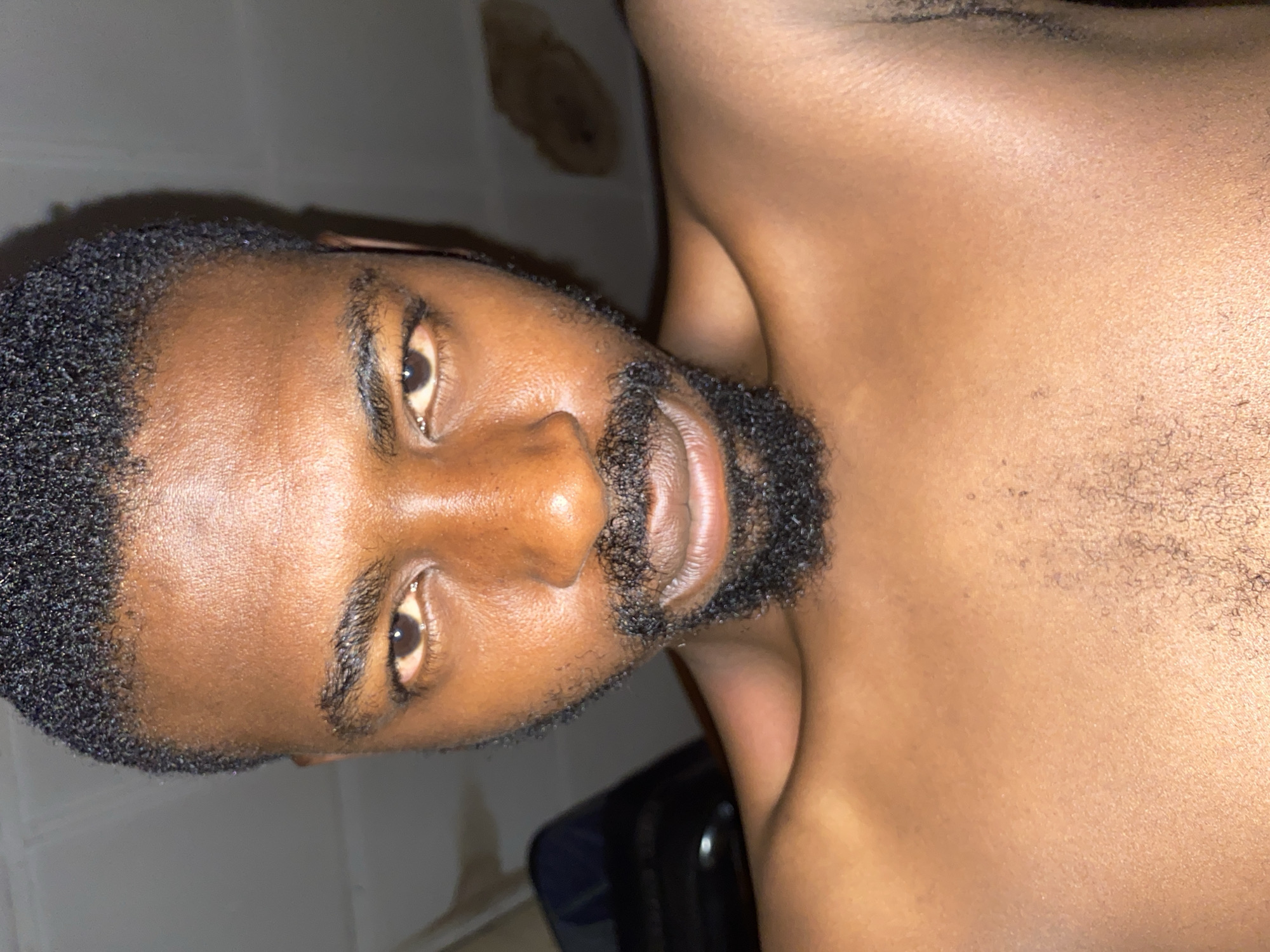 A close-up photograph shows a dark-skinned Black man with short, dark hair and a black beard. His eyes are brown. He is shirtless, and his chest and shoulder are visible. The background appears to be a tiled wall and an unidentified object. Location is unidentifiable as the location in the city of Barakin Akawo, Nigeria.