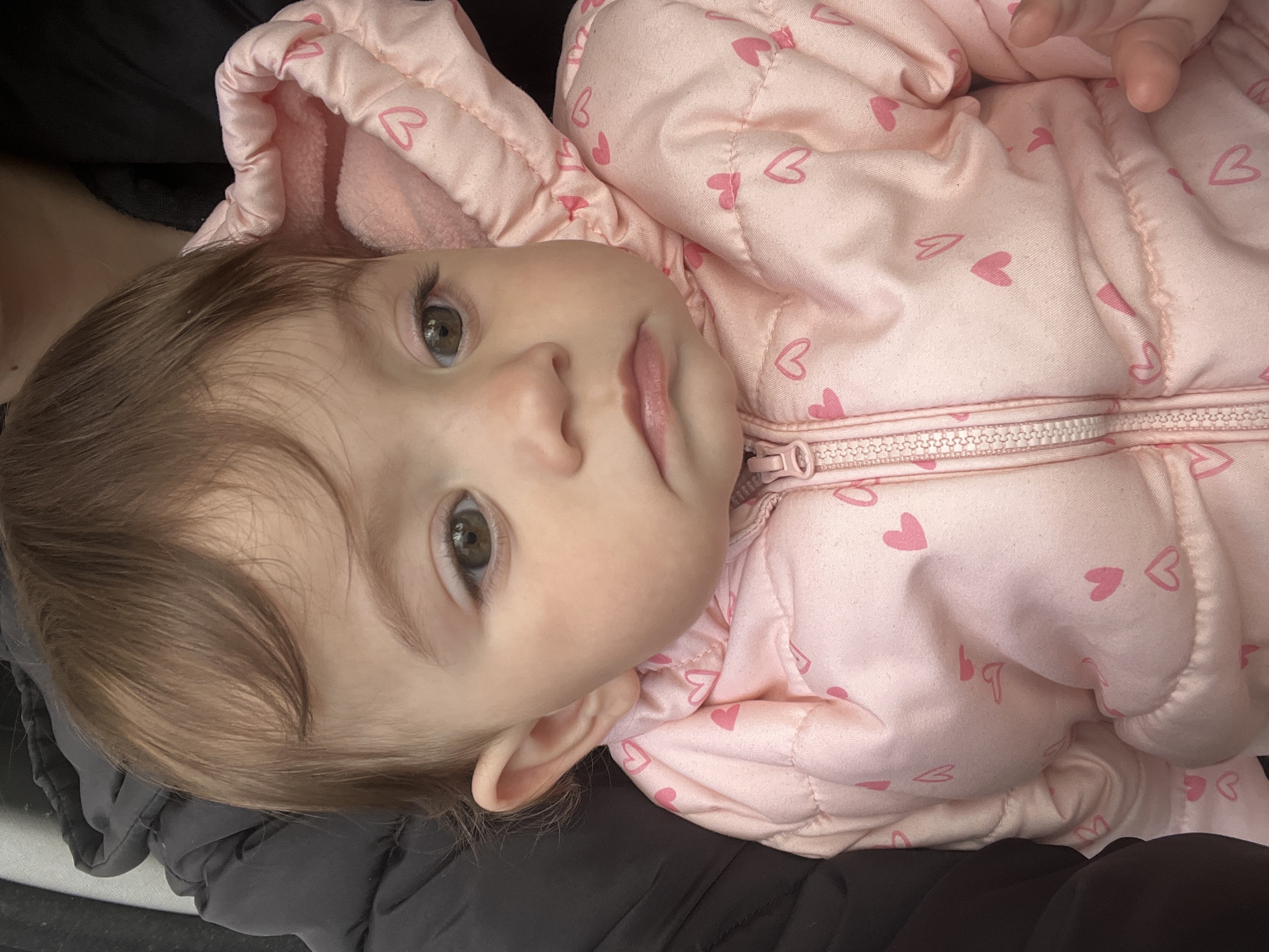 A close-up captures a baby wearing a pink hooded jacket adorned with heart patterns. The child is lying down, with their head slightly tilted towards the viewer. The jacket's zipper is visible near the baby's chest. The baby has fair skin, light brown hair, and light-colored eyes. The background is out of focus and appears to be dark-colored clothing. The scene is not identifiable with a location in the city of Elena, Bulgaria.