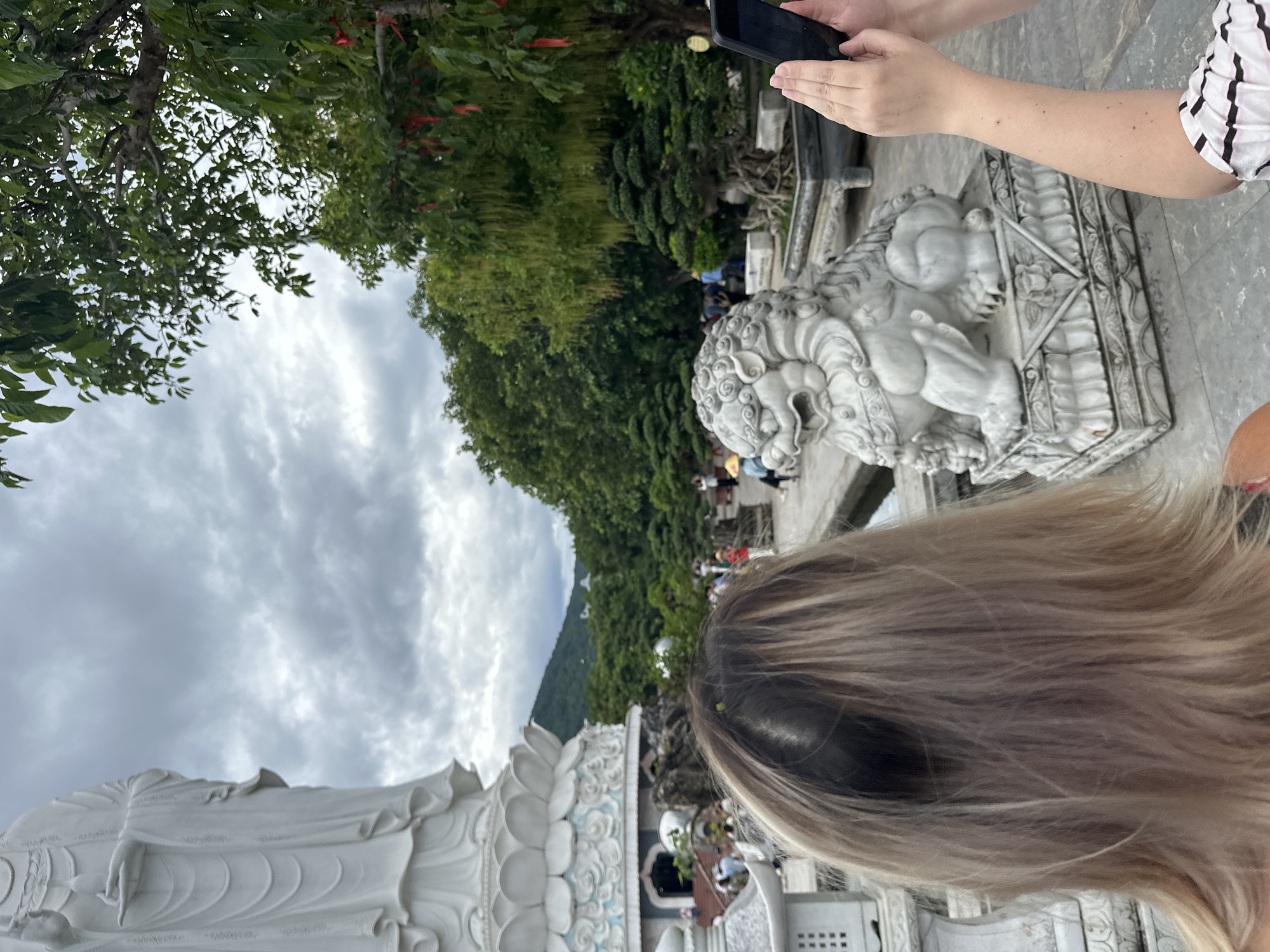 The image shows the Linh Ung Pagoda complex on Son Tra Peninsula in Đà Nẵng, Vietnam. In the foreground, the back of a person with blonde hair is visible, facing a large white statue of a Buddhist deity. To the right of the deity, a white stone statue of a foo dog is displayed on a pedestal. Another person's hands are holding a smartphone, taking a photo of the scene. In the background, there are lush green trees, a cloudy sky, and other visitors to the pagoda. The overall scene depicts a tourist visiting a significant religious and cultural site.