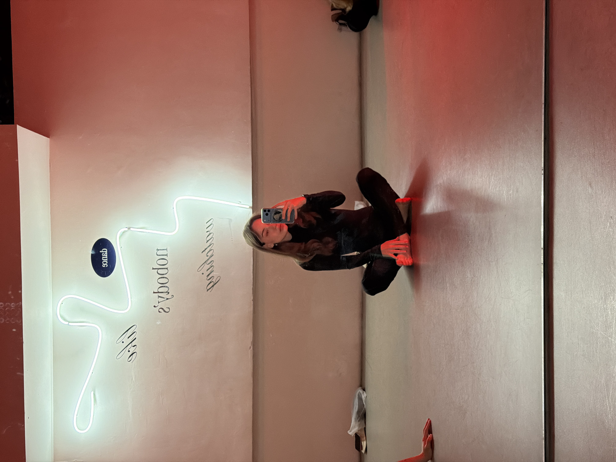 The image displays a mirror selfie of a female individual in an indoor studio environment, illuminated by mixed red and white light.
**Location:** An unspecified studio in Moscow, Russia.
**People:**
A single female subject, with light brown hair, is seated cross-legged on a light-colored floor, facing her reflection. She is dressed in a black long-sleeved top and black trousers. She holds a smartphone with a blue case in her right hand, positioned to capture the mirror reflection. Her left hand rests on her left knee. Her bare feet are visible. A segment of a bare forearm and hand, presumed to be the subject's left arm, is visible resting on the floor in the bottom-left quadrant of the frame, close to the mirror's edge.
**Objects and Environment:**
The room features a large mirror reflecting the entire scene. The wall behind the subject's reflection is a light, possibly off-white or pale pink hue, with an upper section or ceiling corner appearing darker reddish-brown. A white, abstractly shaped neon light fixture is mounted on this wall. English text, reversed due to the mirror reflection, is discernible on the wall: "nobody's perfect", a partial word "life", and "dance" enclosed within a dark oval plaque. The floor is a flat, light-colored (likely grey or tan) hard surface, showing minor scuff marks. The scene is significantly influenced by a strong red ambient light source, which casts a deep red hue on the lower right portion of the floor, the subject's feet, and her right hand. In the background, positioned on the floor near the wall to the left, a white plastic bag and a dark, book-like object are visible. A dark bag or object is also present on the floor at the far right.