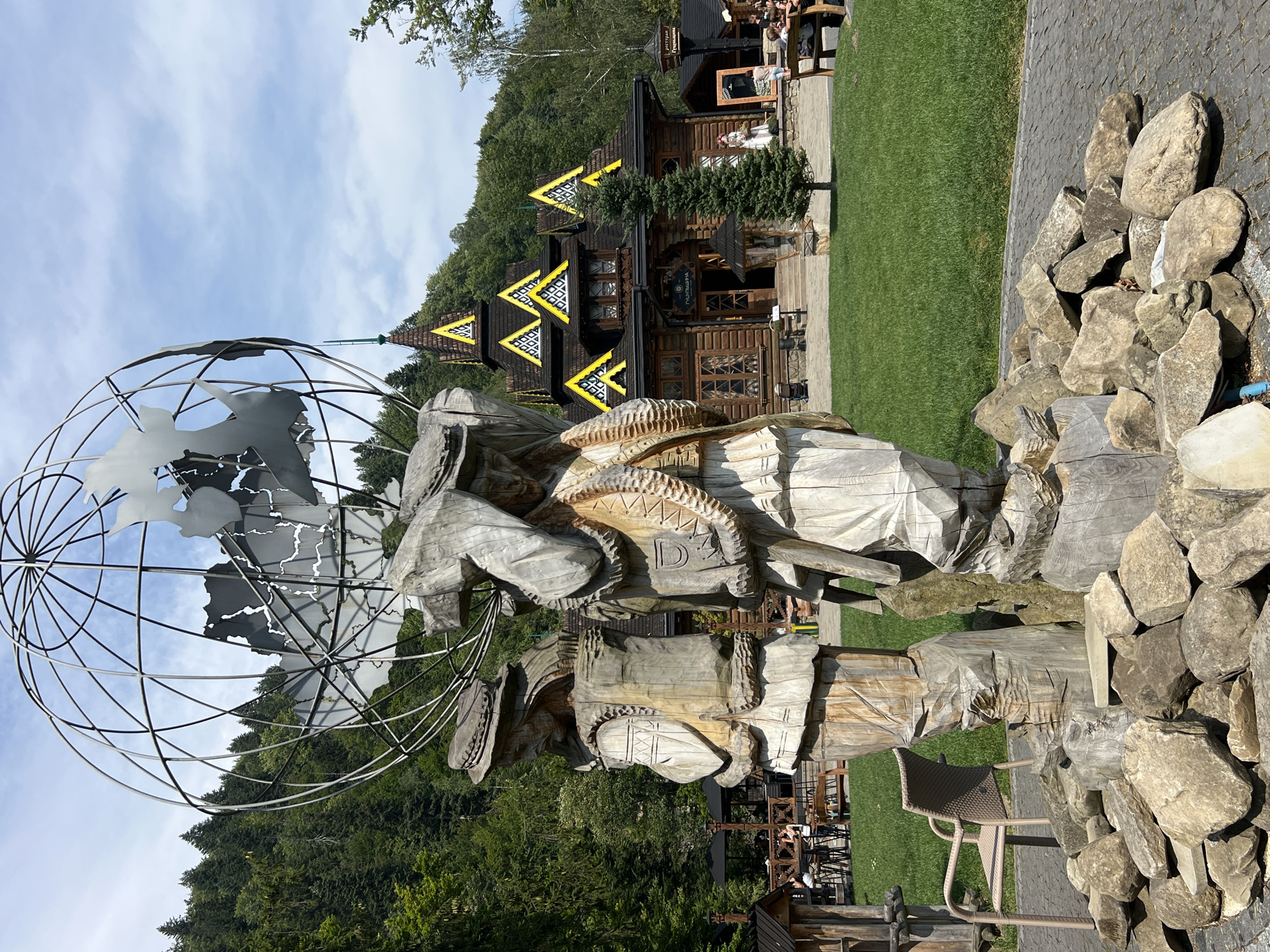 In Yaremche, Ukraine, a large metal globe sculpture with stylized continents stands prominently in front of a rustic wooden building featuring a dark roof with bright yellow triangular accents on its peaks. Two large, intricately carved wooden statues of men in traditional attire stand in the foreground, placed on a base of large stones. The statues depict men wearing hats and vests, one carrying a large object on his shoulders. The ground surrounding the sculptures is paved with cobblestones, and a lush green lawn stretches towards the building. In the background, a dense forest covers rolling hills under a partly cloudy sky. Several people are visible in the distance, some seated at outdoor tables near the building and others on benches. A solitary evergreen tree stands in front of the wooden structure.