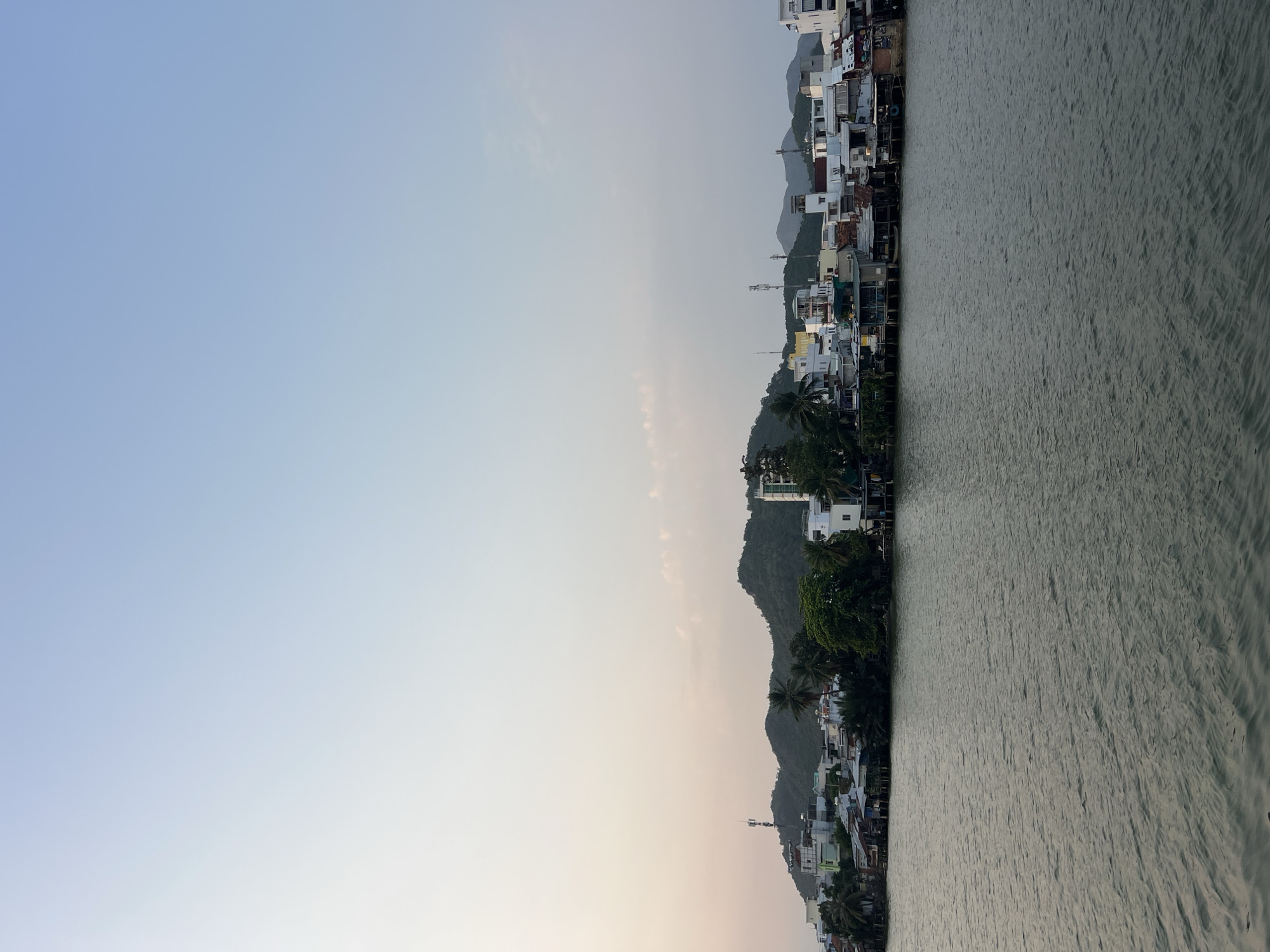 The image depicts a waterfront scene in Nha Trang, Vietnam. The sky is light blue, transitioning to a pale orange near the horizon. A row of buildings, densely packed and varied in height and color, lines the waterfront. Palm trees and other greenery are interspersed among the buildings, with a hilly terrain rising behind them. To the right is a body of water, its surface rippled and gray in color. The image seems to be taken at dusk or dawn.