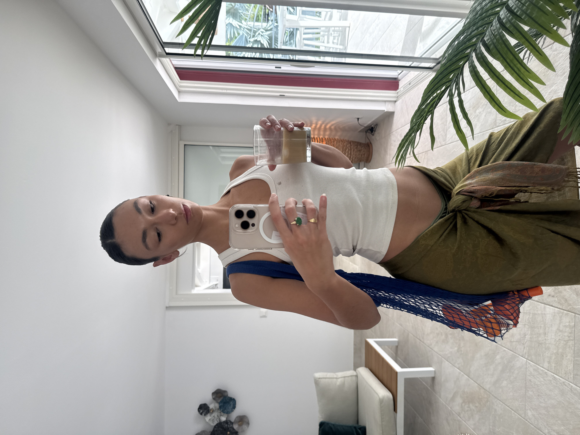 A young person, likely female, is captured in a mirror selfie within a brightly lit, modern indoor setting, presumably in Saint-Martin, France. She has dark hair pulled back and wears a white ribbed tank top paired with olive green trousers that appear to be tied at the waist. She holds her smartphone (an iPhone with a clear case and three cameras) in her right hand, which is positioned to take the photo. A ring with a prominent green stone adorns her right ring finger. In her left hand, she holds a clear glass containing a light brown beverage, possibly coffee or tea. A blue net bag, filled with orange objects that resemble oranges or other fruits, is draped over her left shoulder and arm.
The background features white walls and a light-tiled floor. A large window or skylight structure dominates the upper part of the scene, allowing ample natural light to flood the room. Through this window, glimpses of white buildings with multi-story structures and some greenery are visible, indicative of a warm climate and potentially an urban or semi-urban environment consistent with Saint-Martin. A large, tropical palm-like plant is visible in the upper right corner, contributing to the indoor-outdoor feel. Partially visible in the lower right is a modern white sofa or chair with wooden details. In the lower left, decorative wall art composed of circular, multi-toned elements is displayed. The overall ambiance suggests a casual daytime moment in a contemporary and comfortable home. No visible text is discernible in the image.