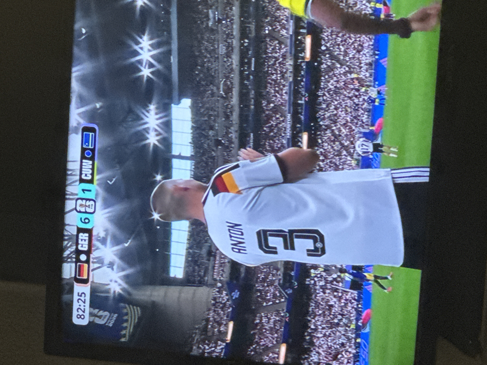 A television screen displays a soccer match. The score is 82:25, with GER on one side and CUW on the other. GER has 6 goals, and CUW has 1 goal. The jersey of a player in the foreground reads "ANTON 3". The crowd in the background is large and appears to be in a stadium. The environment appears to be a soccer field with stadium seating. The location is Unknown, Unknown.