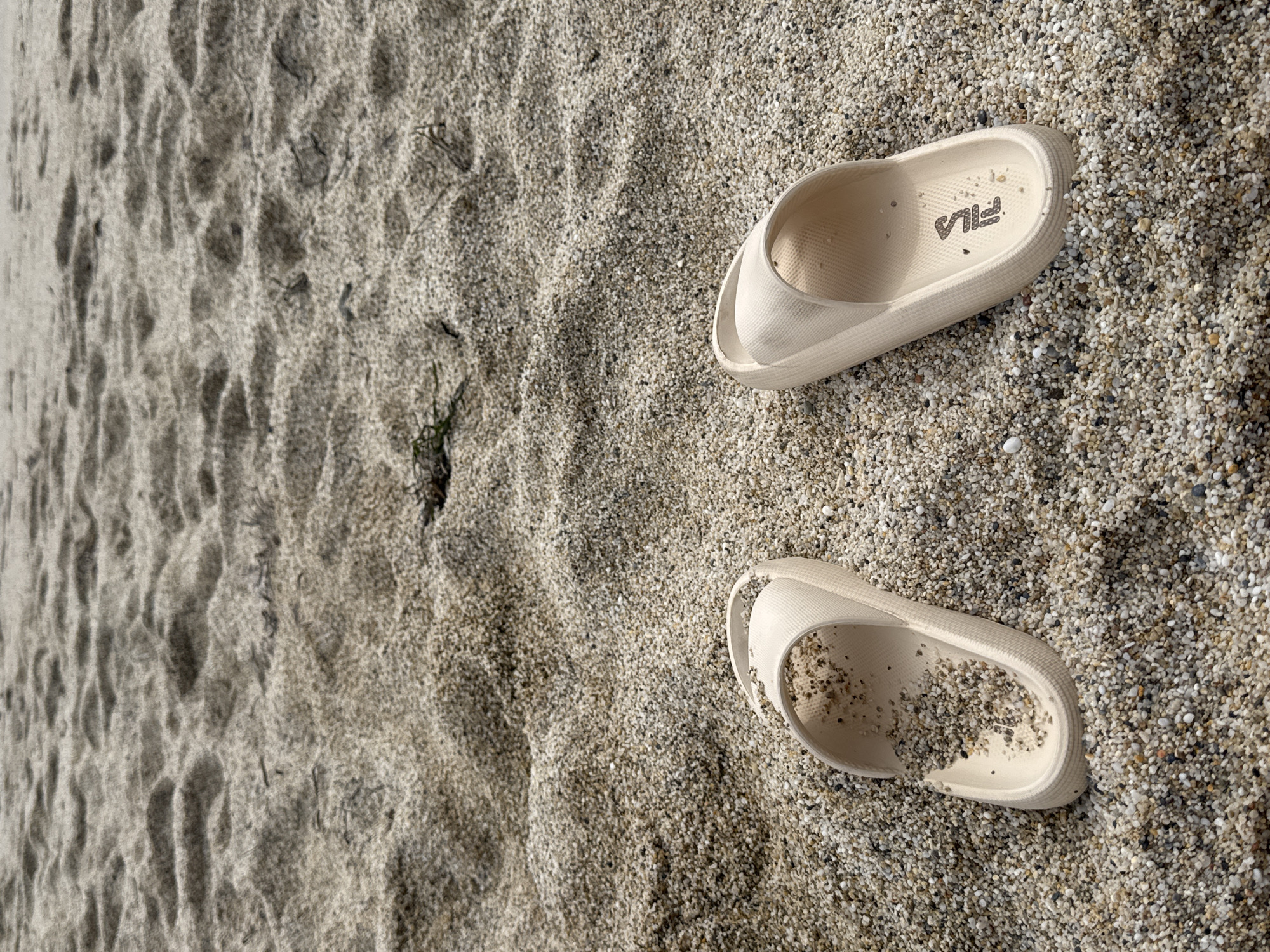 The image shows two beige Fila branded sandals lying on a pebbled beach. The sand contains small pebbles and appears to be slightly damp. One of the sandals contains a small amount of sand inside. The beach has patches of compacted sand and loose pebbles. Given the presence of a pebbled beach in the image and the prompt indicating a location, it is plausible that the photo was taken on a beach in Alanya, Turkey.