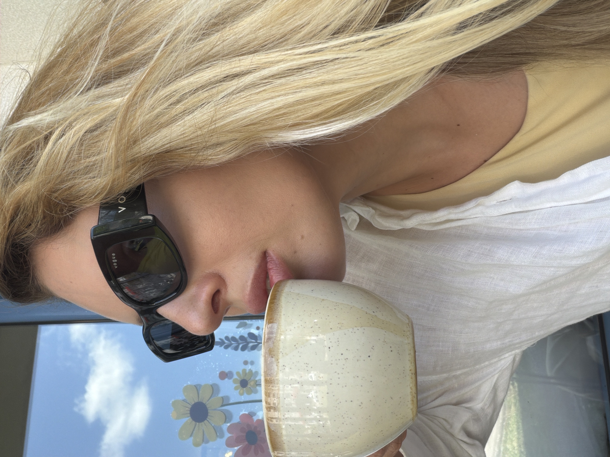 A close-up, profile shot shows a fair-skinned woman with blonde hair, wearing black sunglasses with the "VOGUE" logo visible, sipping from a speckled ceramic mug. She has on a white linen shirt over a yellow tank top. The background appears to be a window with a view of the sky and clouds, and colorful floral decals on the glass. The scene is set outdoors, likely on a sunny day, with hints of greenery visible through the window. The overall impression is one of relaxed enjoyment. The background suggests the location may be a cafe or restaurant with a view, possibly in the city of Budva, Montenegro, known for its scenic coastal setting.