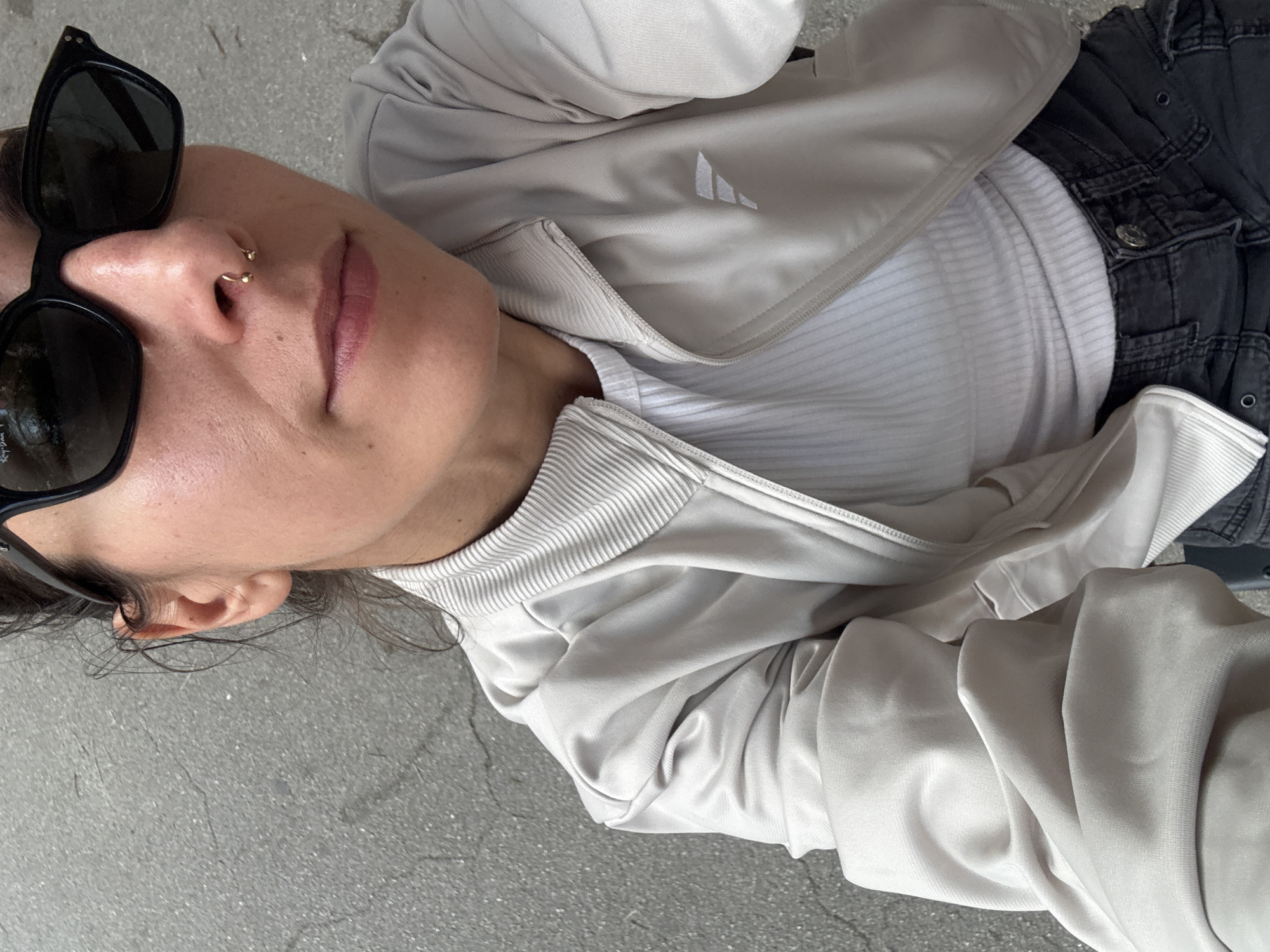 A high-angle close-up selfie captures a young adult female from the chest up, positioned outdoors on a textured grey asphalt or concrete surface in Barcelona, Spain. The individual is wearing dark, rectangular sunglasses with a visible "Ray-Ban" logo on the right lens (viewer's left). A gold horseshoe barbell septum piercing is visible on her nose. She is dressed in a light beige or greige zip-up track jacket featuring a ribbed collar and a small white stylized "F" or similar geometric logo on the left chest (viewer's right). Beneath the jacket, a white ribbed top is visible. Dark grey cargo-style pants with a prominent metal button and grommets at the waistband are also partially seen. Her dark hair is pulled back, with loose strands visible near her right ear. The background shows signs of wear and minor cracks in the pavement, illuminated by bright daylight.