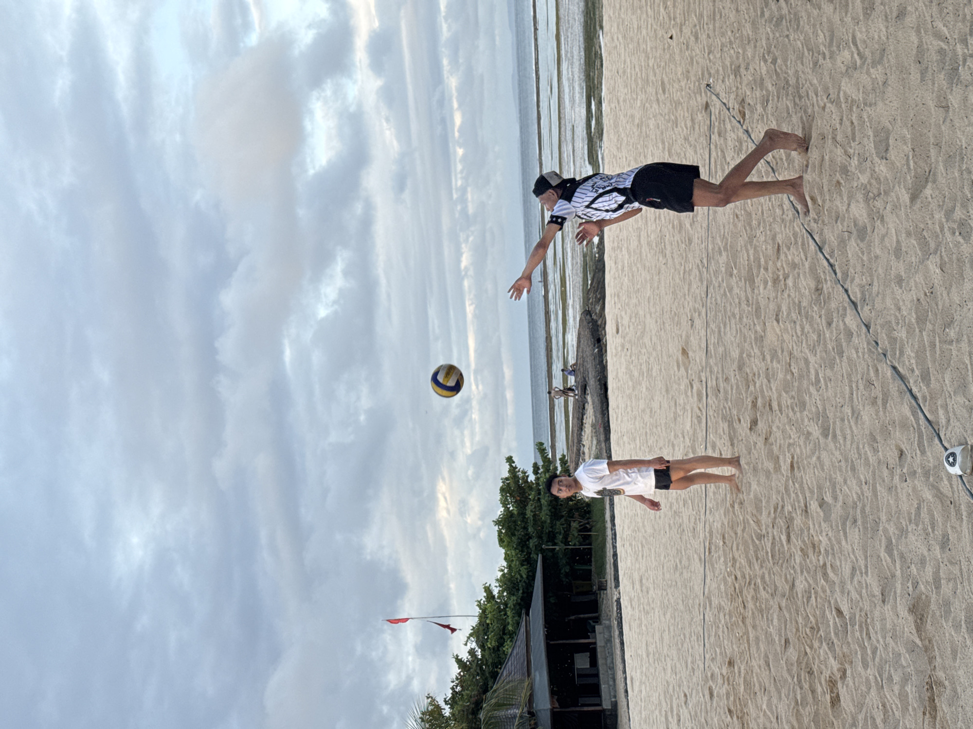 The image captures a beach volleyball scene in Kuta Selatan, Indonesia. A male player wearing a striped shirt, black shorts, and a cap is extending his arm to hit a volleyball. Another male player, dressed in a white shirt and black shorts, stands further back on the sand. A volleyball net stretches between the two players, with a white marker at the net's end. The sand shows footprints and marks indicating prior activity. In the background, a line of vegetation, including trees, and a building with a red flag on top mark the edge of the beach. The sky is cloudy, suggesting the time of day is either early morning or late afternoon. The ocean is visible in the background.