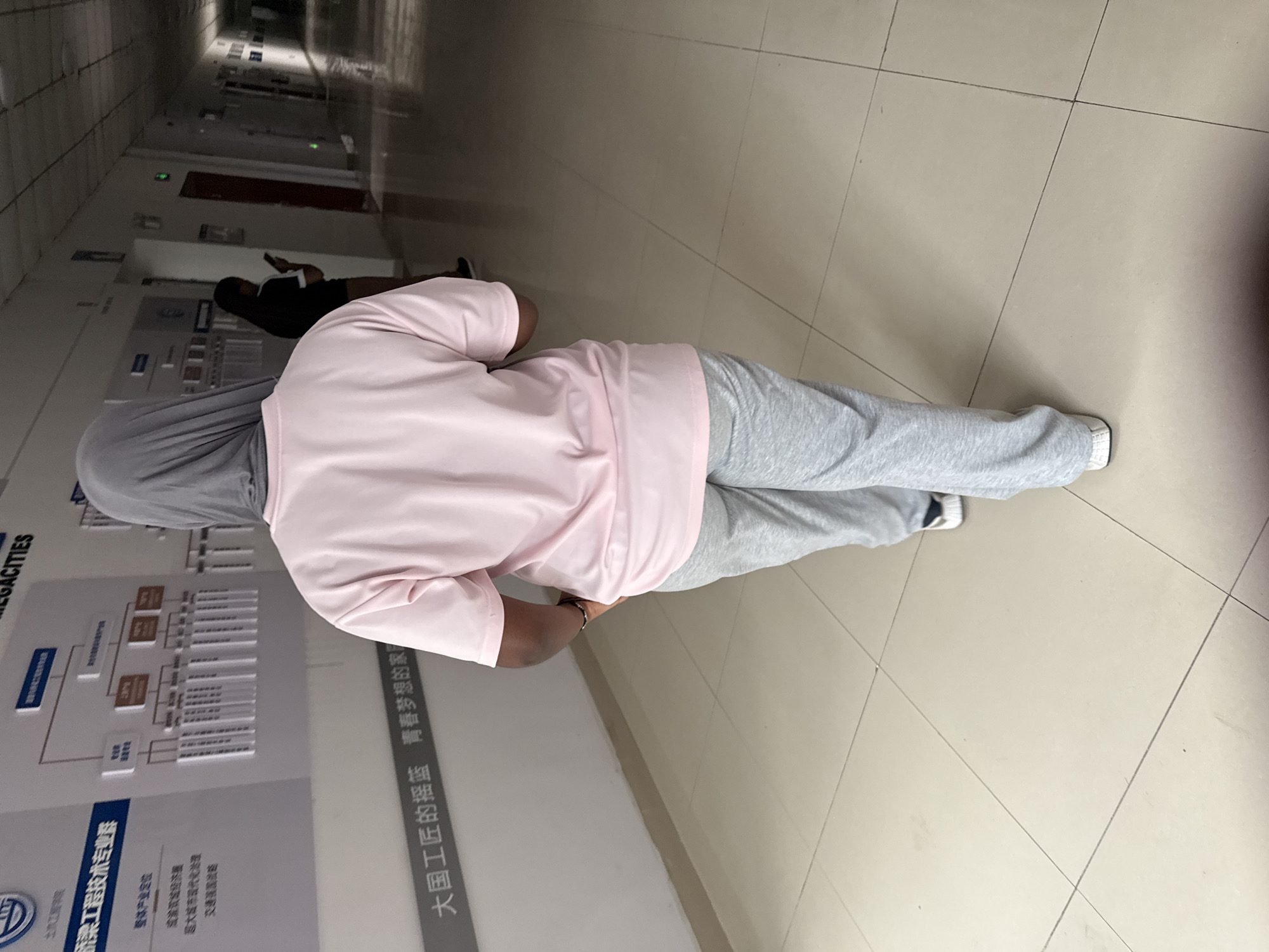 The image shows a person from behind, wearing a light pink t-shirt and grey sweatpants, with a grey hijab. They are standing in a long, well-lit hallway with tiled flooring. On the left wall, there is a large display board with charts and text in Chinese. Another person, wearing a black t-shirt and brown shorts, is visible further down the hallway, looking at their phone. The hallway appears to be part of an institutional building, possibly a school or office. The text on the wall to the left includes "大国工匠的摇篮 青春梦想的家园", which translates to "Cradle of Great Country Craftsmen, Home of Youth Dreams". The presence of this slogan suggests the location is likely in China, and it could be a vocational training center or a similar institution. While the image does not explicitly state it is in Jiangjin, China, the context of Chinese text and the mention of "Great Country Craftsmen" could point towards an educational or industrial facility in China.
