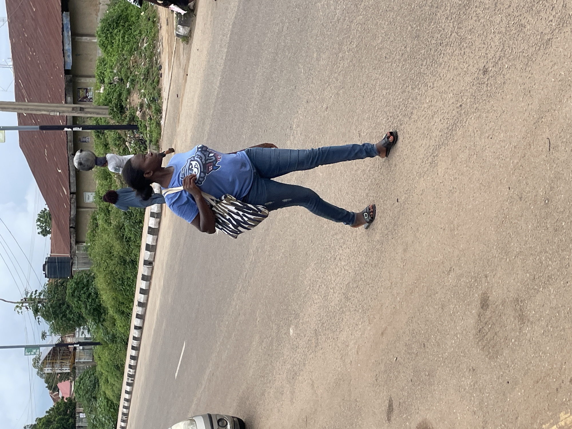 In Jos, Nigeria, a woman walks across a paved road, her blue t-shirt and denim jeans a stark contrast to the muted tones of the surroundings. She carries a patterned bag over her shoulder, her dark hair pulled back. To her left, the partial view of a silver car indicates it is parked or moving on the road. The road itself is worn and dusty, lined with a black and white striped curb that separates it from a grassy verge. Beyond the verge, a collection of buildings with corrugated iron roofs and cinder block walls are visible, some partially obscured by lush green foliage. A black water tank stands prominently near one of the buildings. In the background, other individuals are seen, one carrying a large spherical object on their head, and another walking away from the camera. The sky above is overcast, suggesting a cloudy day.