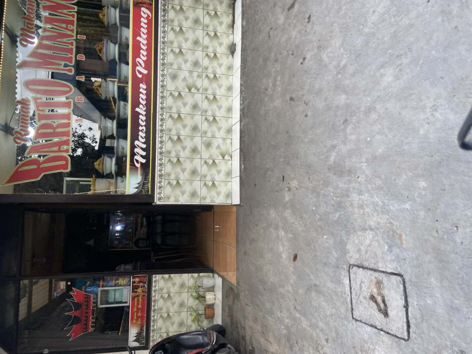 The image shows the exterior of a storefront in Kecamatan Cimahi Selatan, Indonesia. The storefront is marked with a sign that reads "Rumah Makan Barito Minan" in red lettering with the words "Masakan Padang" in black lettering on a red background below. The storefront is made of dark brown wood, with a glass window display. The window display is filled with various food items, including plates of food and bowls of food. There are two potted plants in front of the store, and a motorcycle parked in the foreground. The ground in front of the store is made of concrete. The motorcycle is facing the camera and is partially obscured by the right edge of the image. The ground is light grey, with a drainage grate. The front of the building is tiled in an alternating pattern of white, grey, and tan. The door to the store is open, and the interior of the store is visible. The interior has a grey and white tiled floor, two stools, and a couple of tables. The location of the store can be determined by the coordinates provided: -6.932, 107.564. The scene appears to be abandoned or unoccupied. No people are present, and there are no other signs of activity.