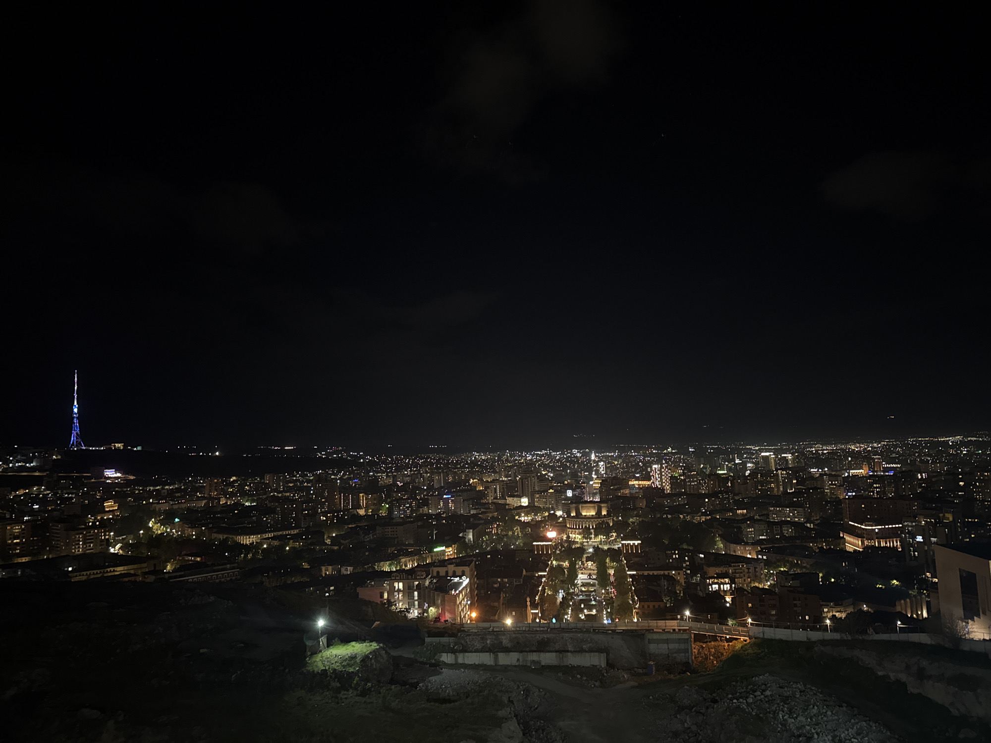 The image depicts a night cityscape, identified as Yerevan, Armenia, by the presence of the Yerevan TV Tower. The view is captured from an elevated position overlooking the illuminated city below. The foreground consists of a dark, uneven terrain, possibly a hillside or a partially constructed area with exposed soil and some vegetation. In the middle ground, city structures and streets are visible, illuminated by artificial lights, indicating a developed urban area. Buildings vary in height and architecture, suggesting a mix of residential and commercial constructions. The background is dominated by the night sky, which is predominantly dark with some cloud cover. The Yerevan TV Tower, a tall, distinctive structure, is located on the left side of the horizon, adding a specific geographic marker. The overall atmosphere is that of a quiet night in an urban setting.