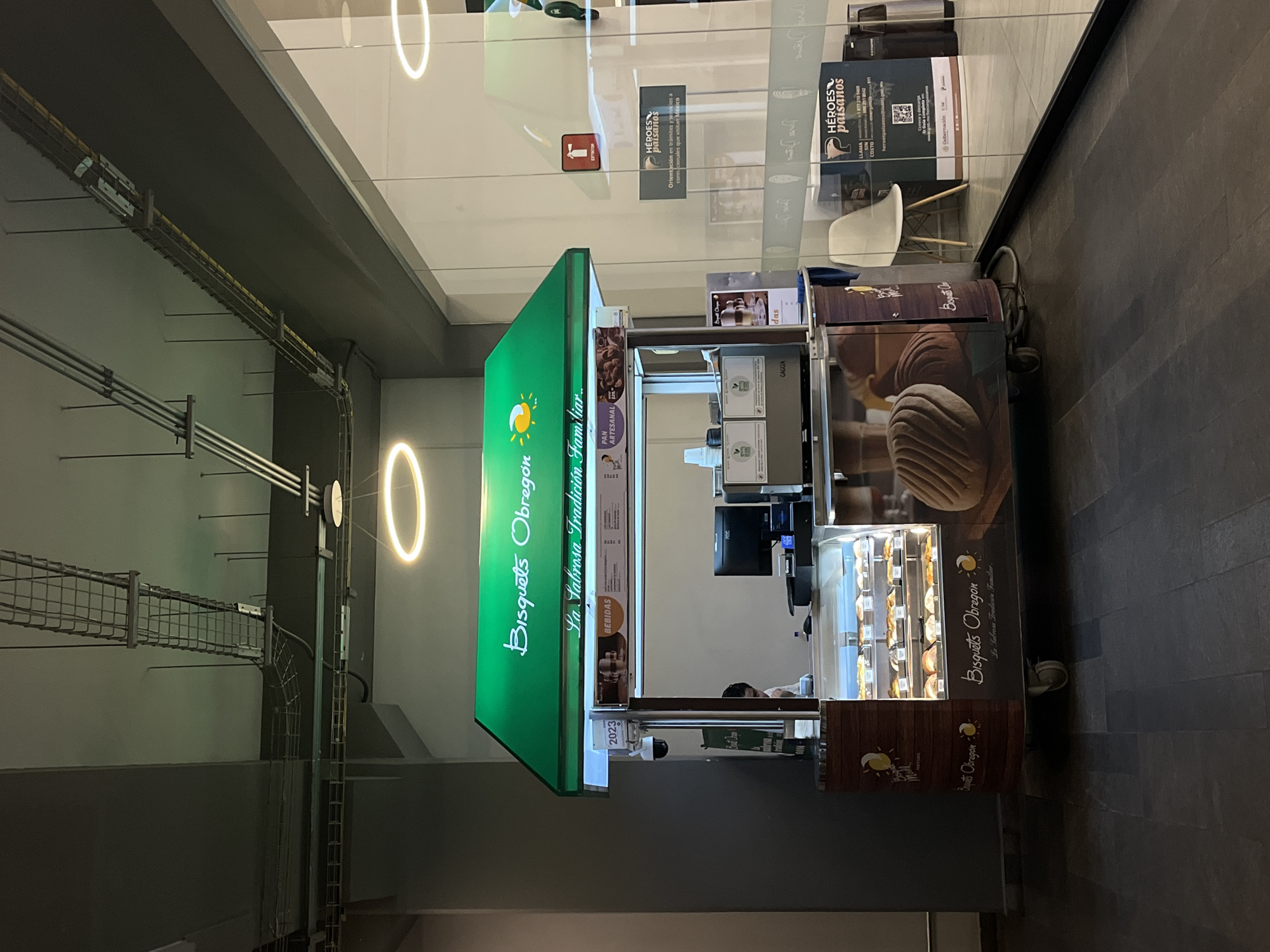 A brightly lit food kiosk stands inside a modern building with polished floors. The kiosk, labeled "Bisquets Obregon," features a green awning with the brand name and a tagline in Spanish. Inside the kiosk, there are food displays showcasing pastries. The kiosk includes equipment like a coffee machine and a point-of-sale system. Signage about "Heroes Paisanos" is visible on the glass wall behind the kiosk, providing information for visiting Mexican nationals. The surroundings consist of a corridor with exposed ceiling infrastructure and circular lighting fixtures. The image conveys the commercial environment of a shopping mall or similar location, possibly within Puebla, Mexico, given the reference to "Bisquets Obregon" and the "Heroes Paisanos" program.