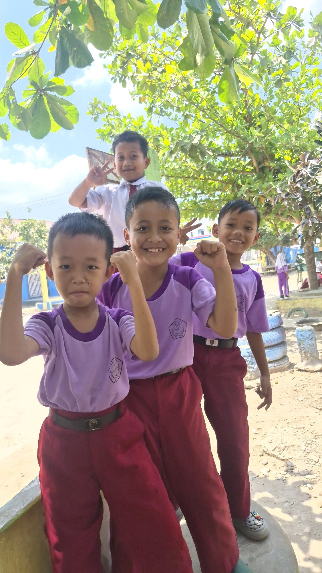 Four young boys, appearing to be of elementary school age, are posing for a photograph outdoors in Putak, Indonesia. The boys are wearing school uniforms consisting of light purple shirts with emblems and dark red pants. Three of the boys in the foreground are smiling and striking poses, with two of them flexing their biceps. Another boy is partially visible behind them, also smiling and making a peace sign. The background suggests a schoolyard or outdoor recreational area with trees, foliage, a blue fence, and some stacked tires. The lighting indicates it is daytime, with sunlight filtering through the trees.