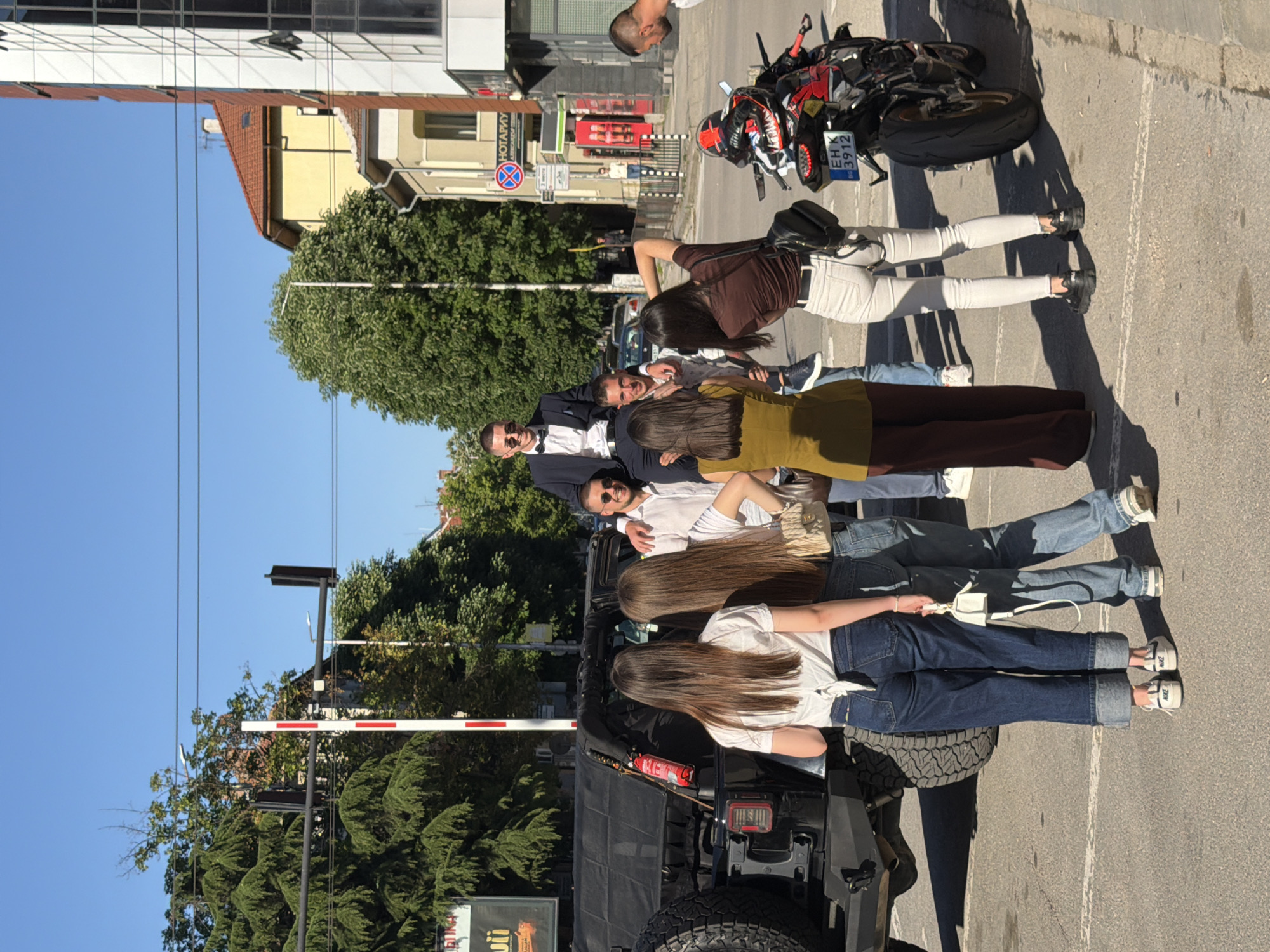 The image depicts a group of people posing for a photo in an outdoor setting. A black Jeep-style vehicle is parked on the pavement, with several individuals standing behind and around it. The group includes women in white shirts and jeans, and men dressed in casual and formal attire. A motorcycle is parked nearby. In the background, buildings and trees suggest an urban environment. A road sign with a European-style symbol is visible. Based on the license plate "EH" and the road sign, the image is likely taken in Pleven, Bulgaria.