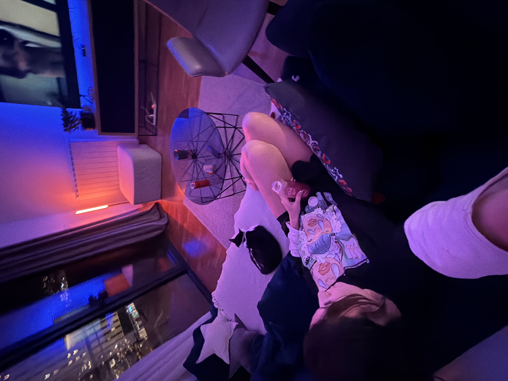 A person is seen in a dimly lit room. The subject is wearing a black shirt and leggings with a colorful, cartoon-style graphic design. A white pillow is visible next to the person. An end table with a metal frame and glass top sits nearby. A city view can be seen through the window in the background, with city lights illuminating the night sky. The city is identified as Belgrade, Serbia.