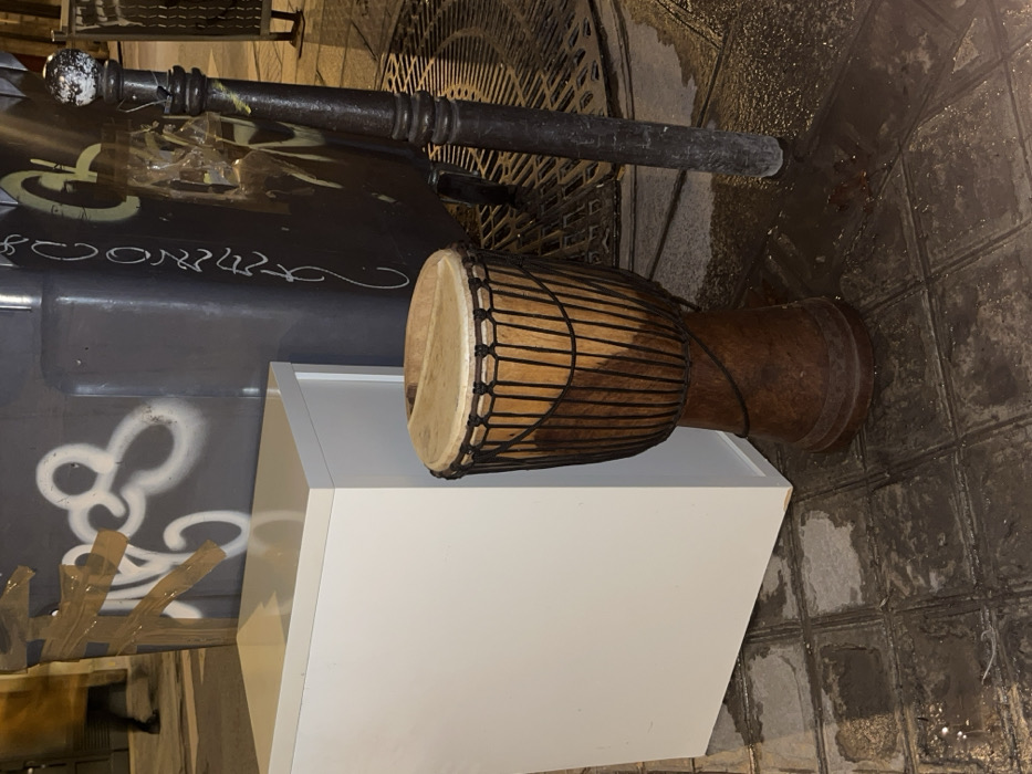 A djembe drum rests on top of a white, cubic structure. The drum has a light-colored drumhead and a dark brown body, secured with black rope. The structure is simple and unadorned. The scene appears to be outdoors, possibly on a city street with wet, tiled paving. Part of a black, cylindrical object with a ball-shaped end is visible in the background. Graffiti decorates a surface to the left.