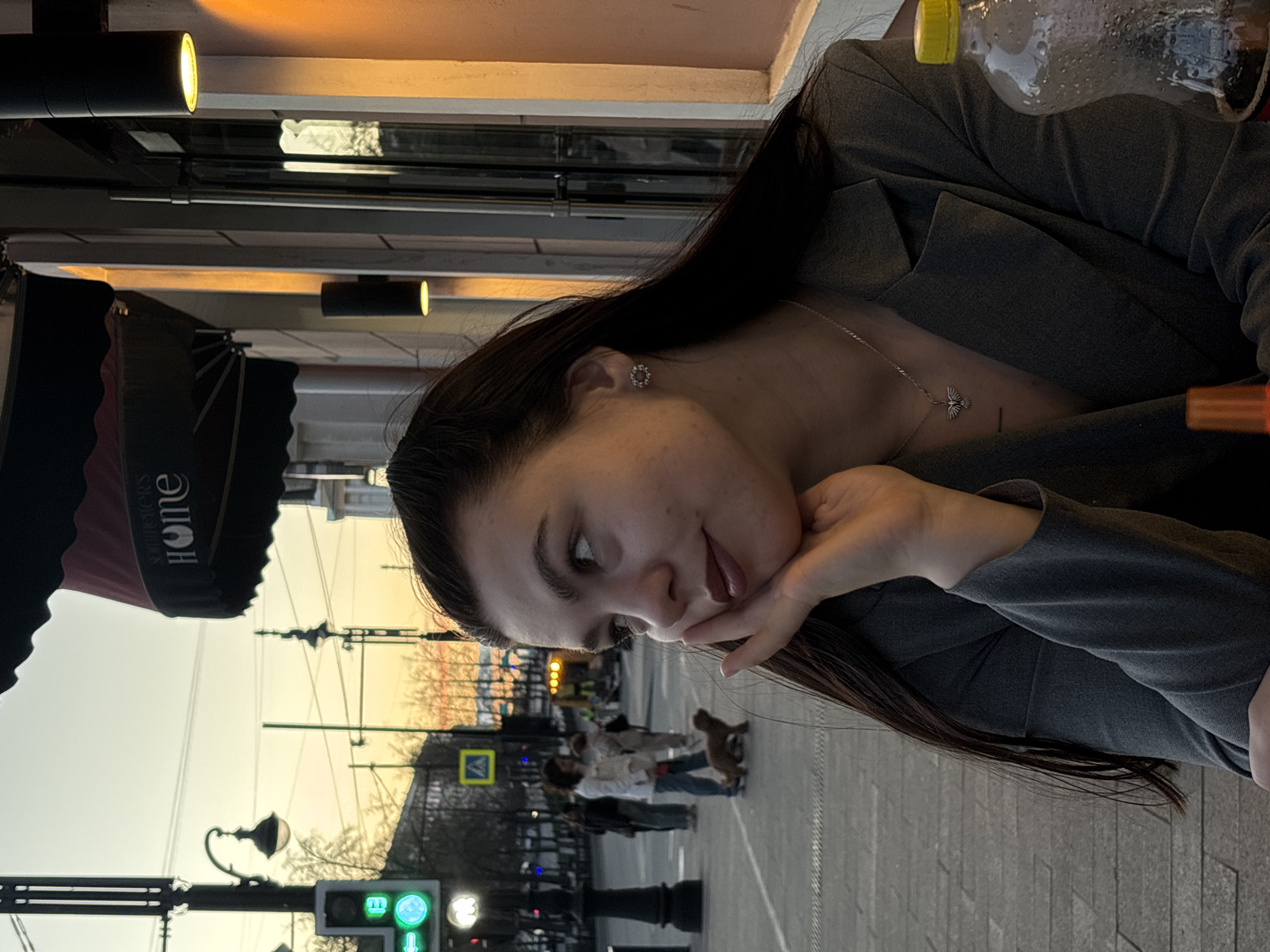 A young woman with long dark hair, wearing a gray jacket and a necklace with a pendant, is seated at an outdoor cafe. Her head is resting on her hand. In the background, a street scene in Perm, Russia, is visible. People are walking on the sidewalk, and traffic signals are present. Streetlights and buildings line the street.
