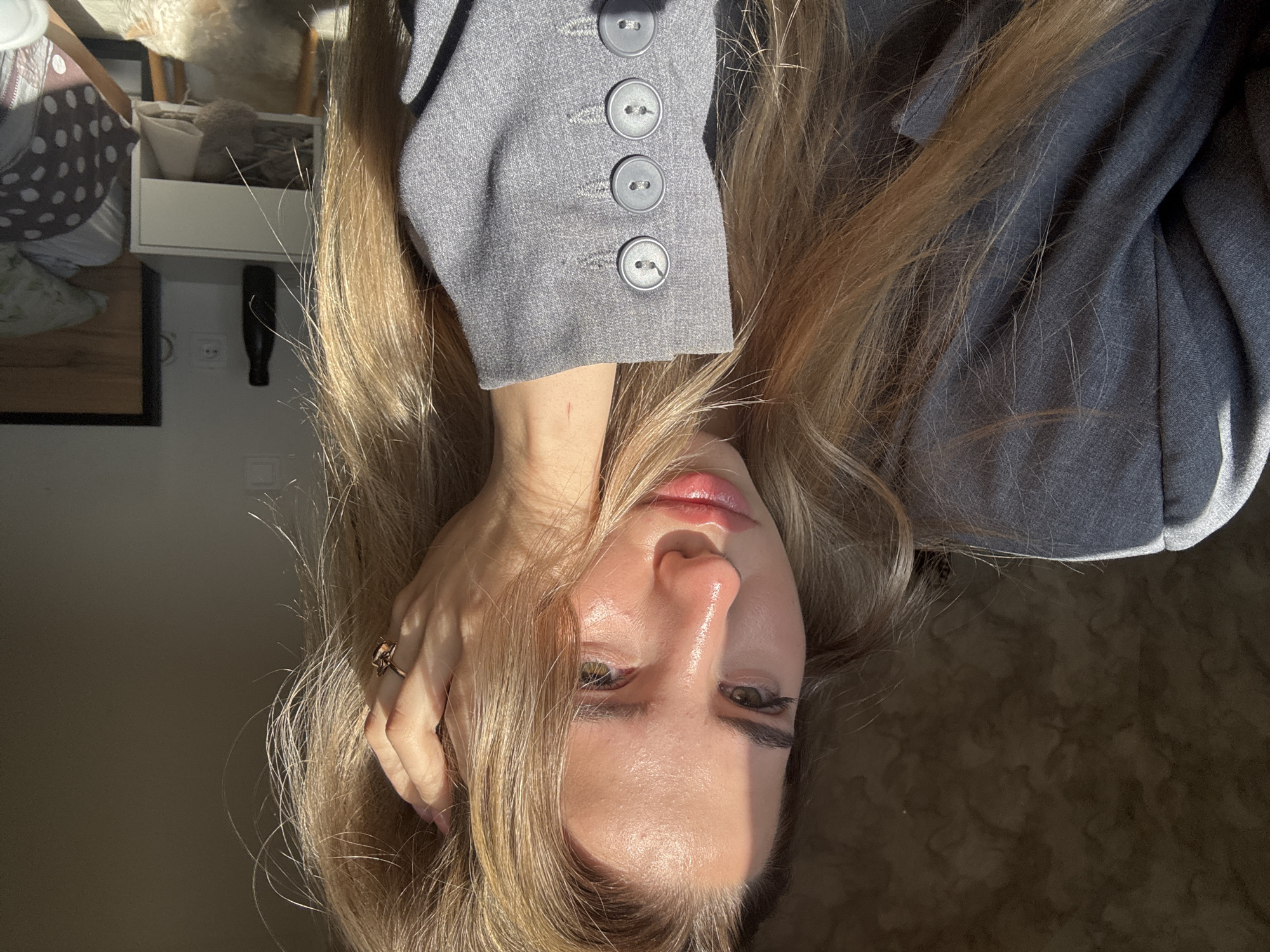 This media file presents a close-up portrait of a person, likely female, with long, light blonde hair, captured indoors in Noviy, Russia. The image is oriented as if the subject is lying down on their side, looking towards the camera, with the frame rotated 90 degrees clockwise.
Strong, natural light, likely sunlight, illuminates the scene, casting bright highlights on the subject's forehead, nose, and lips, suggesting a dewy complexion. Her expression appears calm and serene. She is wearing a grey blazer or jacket, the sleeve of which is prominently featured on the left side, showcasing four large grey buttons. A ring is visible on her right hand, which gently touches her hair near her head, allowing some strands to fall across her face.
In the background, details of a domestic setting are visible. To the original top-left, there is a white floating shelf or small cabinet containing various soft items, possibly textiles. Below it, a power outlet is visible on a wall, with a dark, slender object hanging from it. Further left, a patterned fabric or pillow with white polka dots on a grey base can be seen. The background on the original right side of the image consists of a dark, textured surface, possibly a carpet or bedding. There is no visible text in the image.