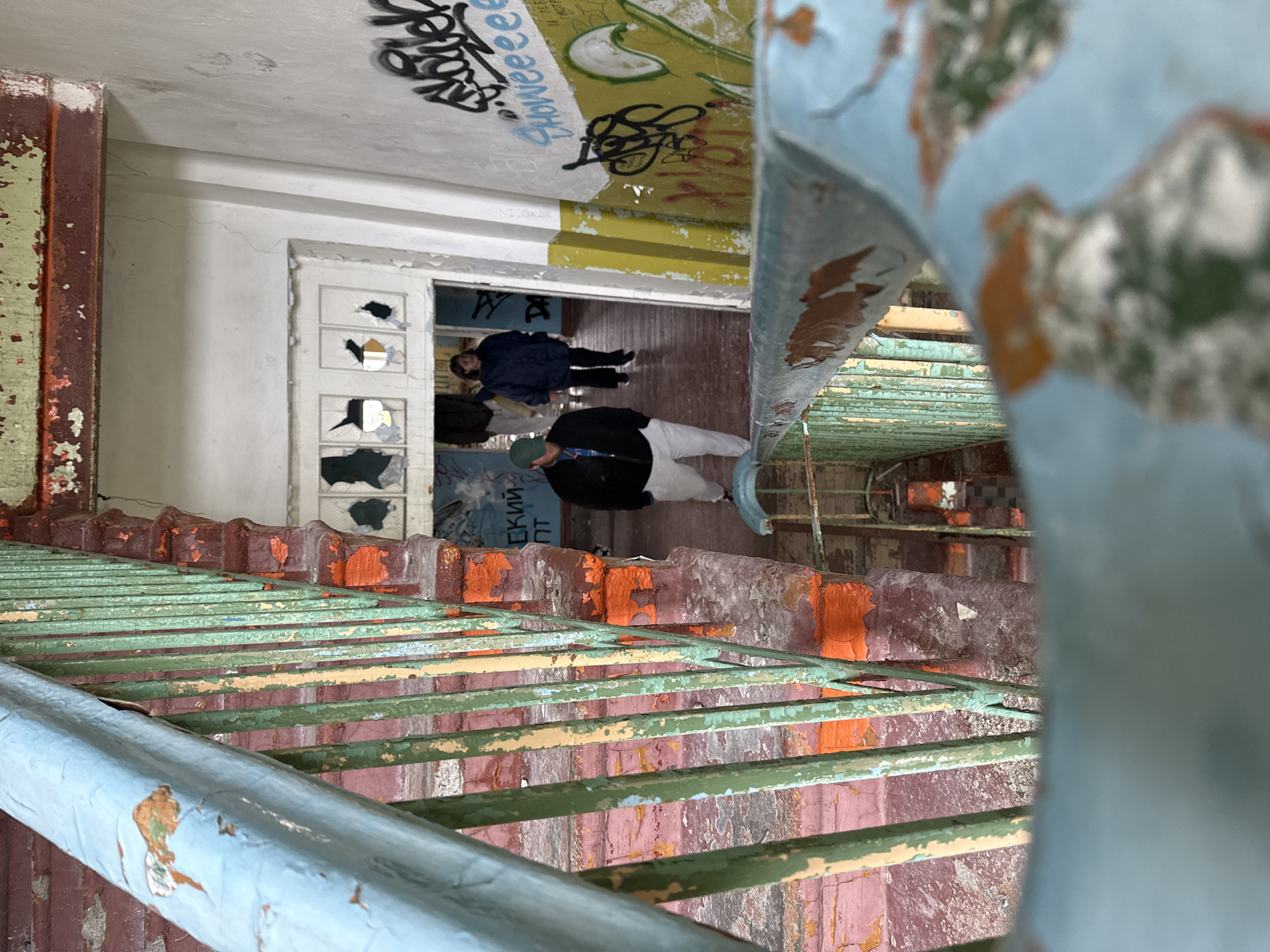 The image depicts a dilapidated interior staircase with peeling paint in an abandoned building in Teriberka, Russia. Two individuals are visible further down a hallway leading away from the staircase. One person, wearing a green cap, black jacket, and white pants, is walking towards the camera. Another individual, wearing a dark blue jacket and black pants, is walking away from the camera. The walls are covered in graffiti, and the overall scene conveys a sense of decay and neglect.