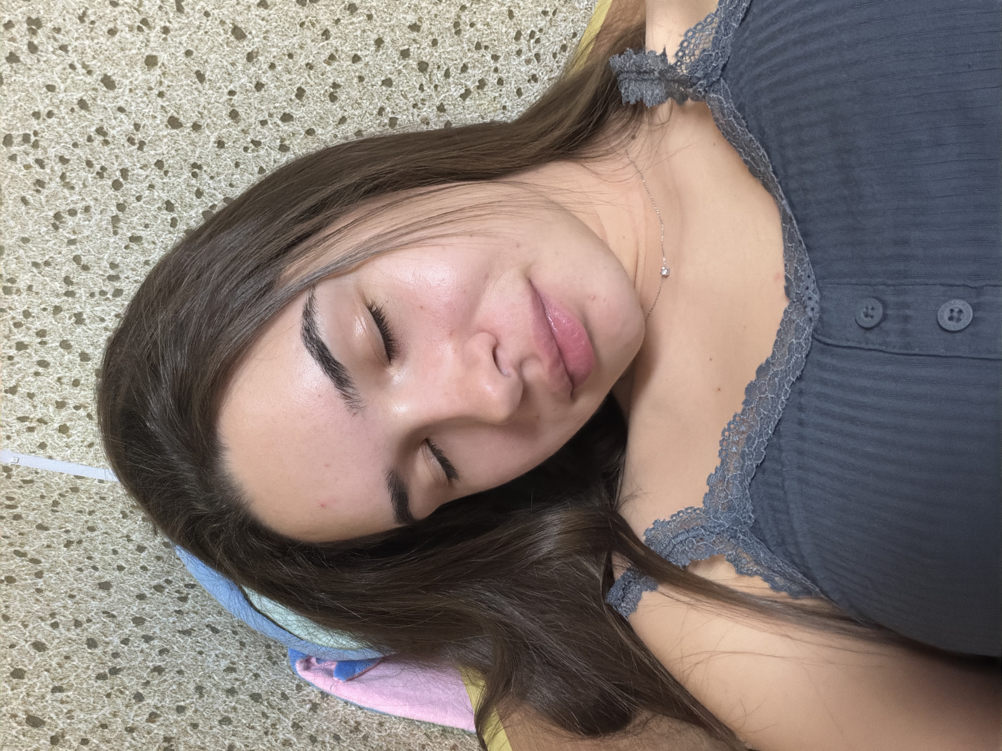 A young woman lies with her eyes closed, resting on a bed or similar soft surface, in an interior setting. She has long, dark brown hair and is wearing a grey top with lace trim. A delicate necklace with a single gemstone is visible. The surface she is lying on has a beige textured pattern. A pillow with muted colors is also partially visible beneath her head. The image does not provide any explicit information to determine the location in the city of Sofia, Bulgaria.