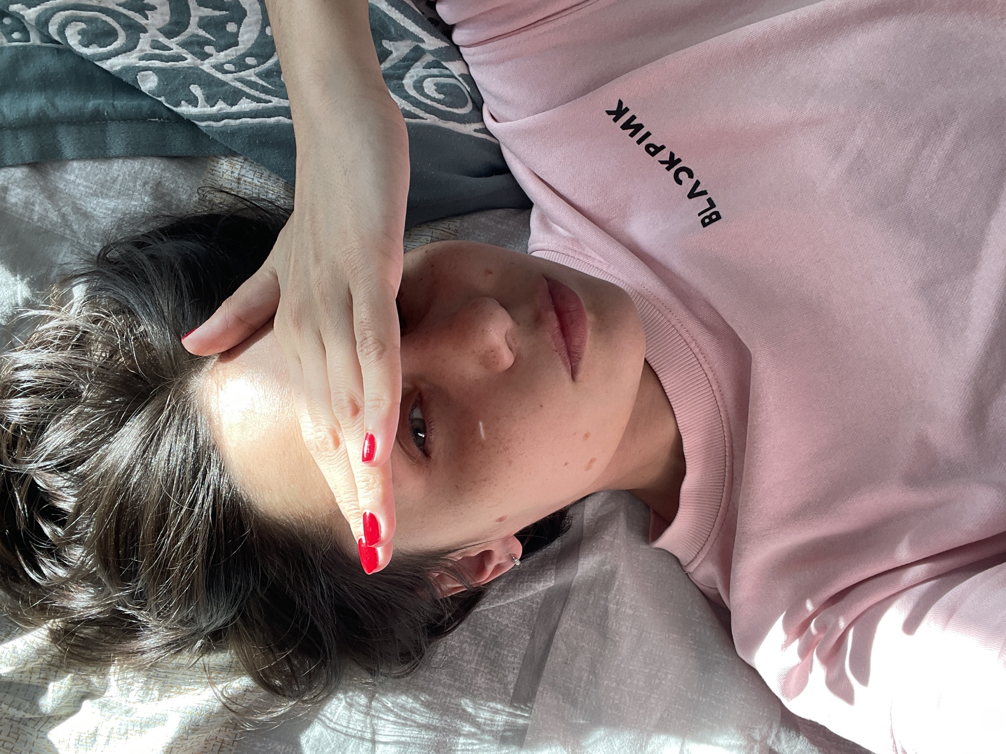 A woman lies on a bed, partially covering her face with her hand. She has dark hair and fair skin with freckles. Her fingernails are painted red. She wears a light pink sweatshirt with the word "BLACKPINK" printed in black. The bed is covered with a patterned gray and white blanket and a darker patterned fabric partially visible at the top of the frame. Sunlight illuminates parts of the scene. There is no evidence in the image to suggest a location in Izhevsk, Russia.
