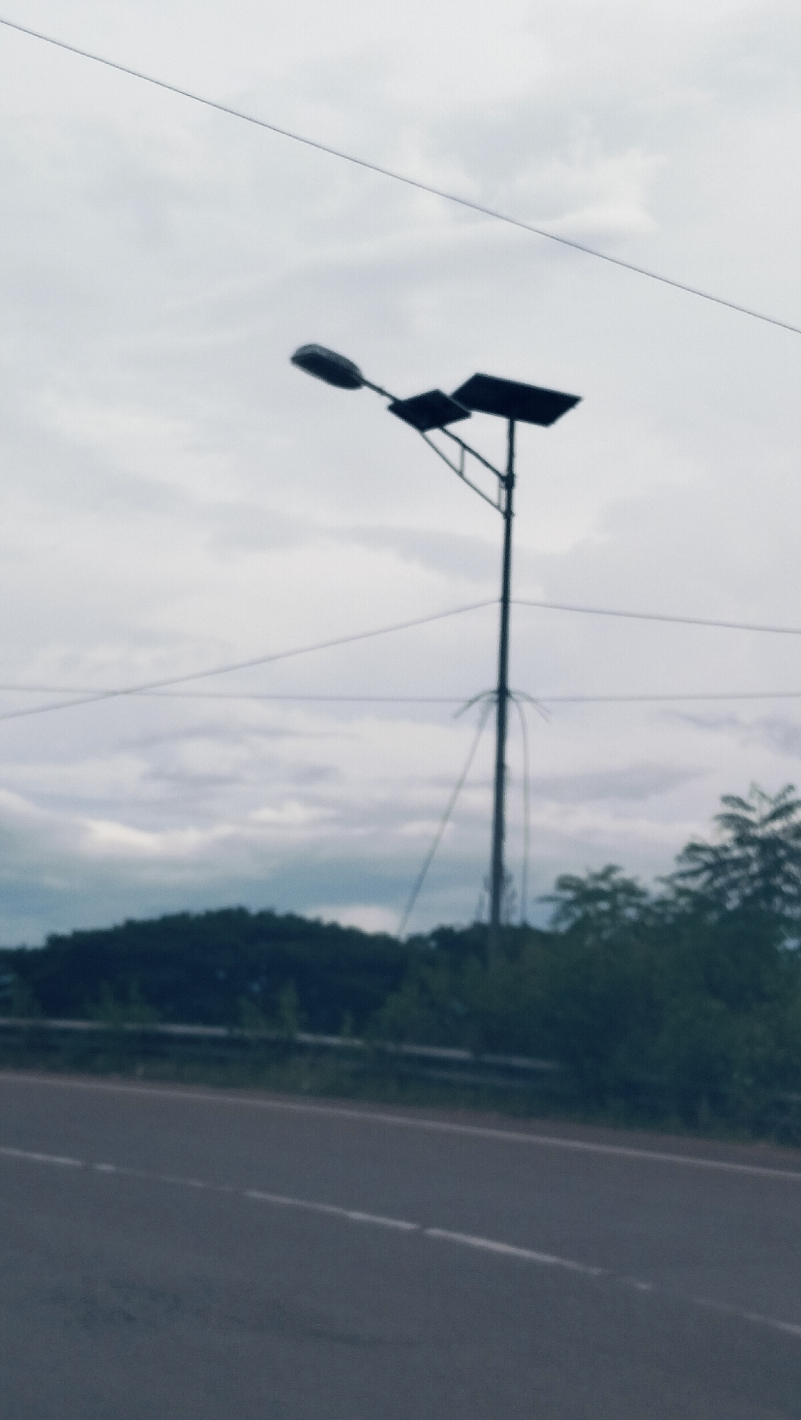 The image depicts an outdoor roadside scene in Lonbaroh, Indonesia, on an overcast day. The primary subject is a solar-powered street light, centrally positioned. It features a tall, dark pole with a single light fixture extending to the left and a rectangular solar panel mounted to the right. Several thin, dark lines, likely utility wires or cables, are visible crossing the muted, cloudy sky.
In the mid-ground, a dense stretch of dark green trees forms a natural horizon, possibly indicating a hill or forested area. A dark, paved road with white dashed lane markings occupies the foreground, running horizontally across the bottom of the frame. A silver-colored guardrail is partially visible between the road and the treeline.
The sky is uniformly grey with soft cloud formations, suggesting a cloudy or heavily overcast weather condition. No specific time of day is discernible, though it is clearly daylight. There are no people, vehicles, or explicit activities taking place, presenting a quiet, static view of the roadside environment. No visible text is present in the image.